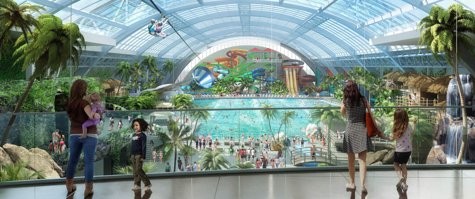 Walkway with glass bannister looks out over waterpark with plant lined walls and water slides at far end