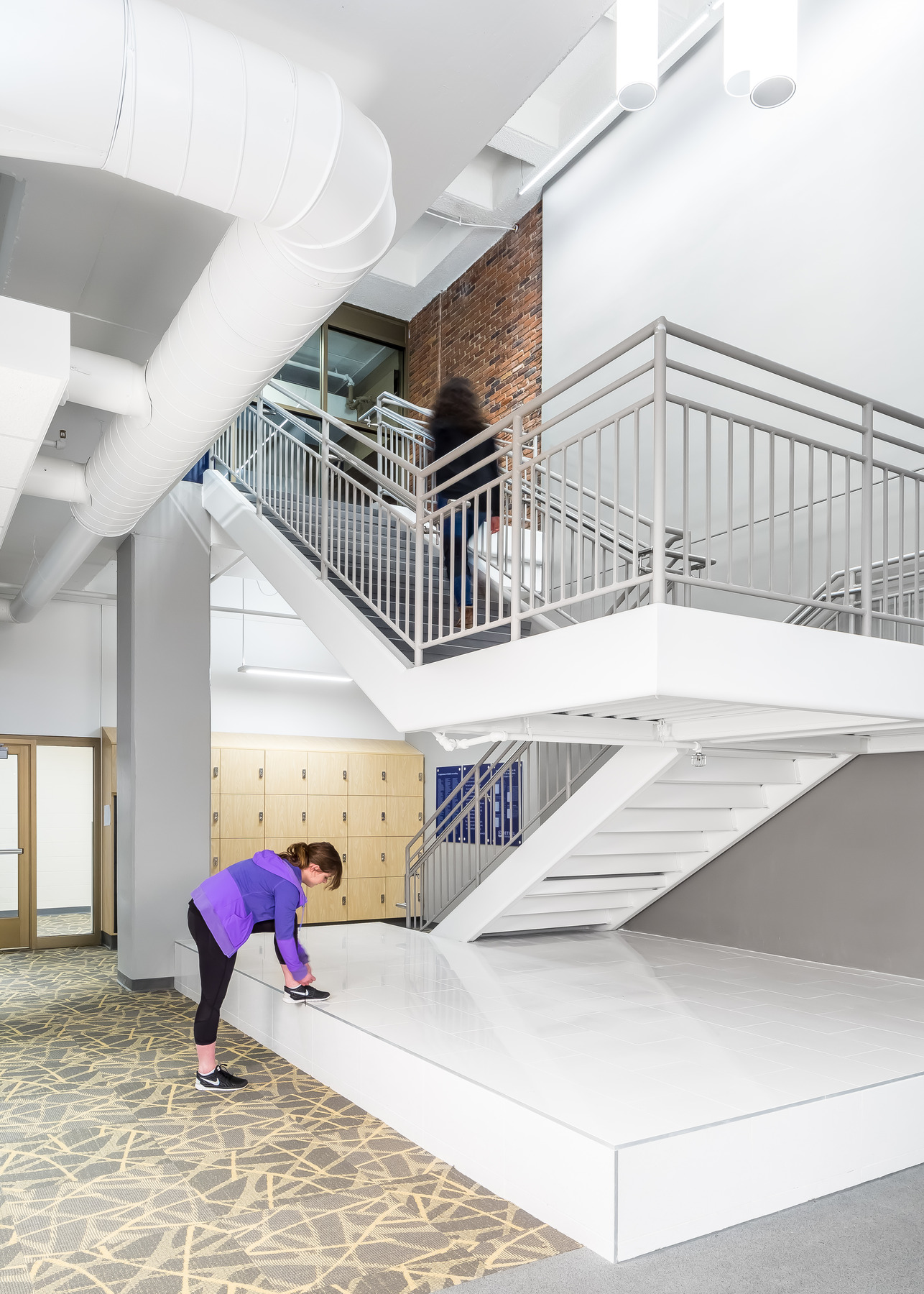Bright, modern interior with a wide white staircase and metal railings in an open lobby. Exposed white ductwork and a brick accent wall rise to a high ceiling.