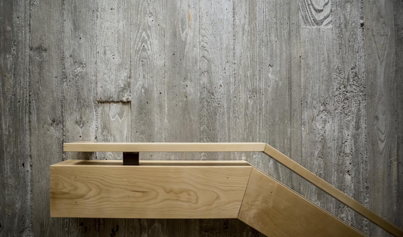 Textural grey raw concrete wall with blonde wood railing angled from right, flattening at center Textural grey raw concrete wall with blonde wood railing angled from right, flattening at center
