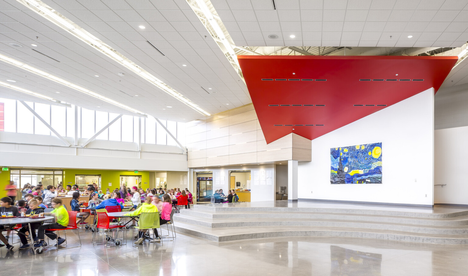 Double height cafeteria with angled red accent connecting with wood paneled wall. Van Gogh's Starry Night hangs by platform