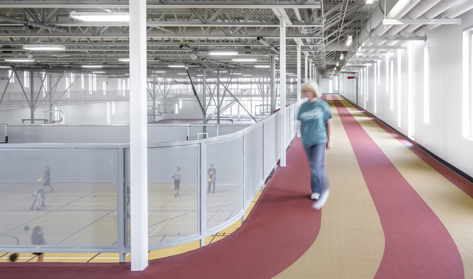 Striped 2nd story track overlooks gymnasium with basketball courts. Exposed beams above with basketball hoops attached