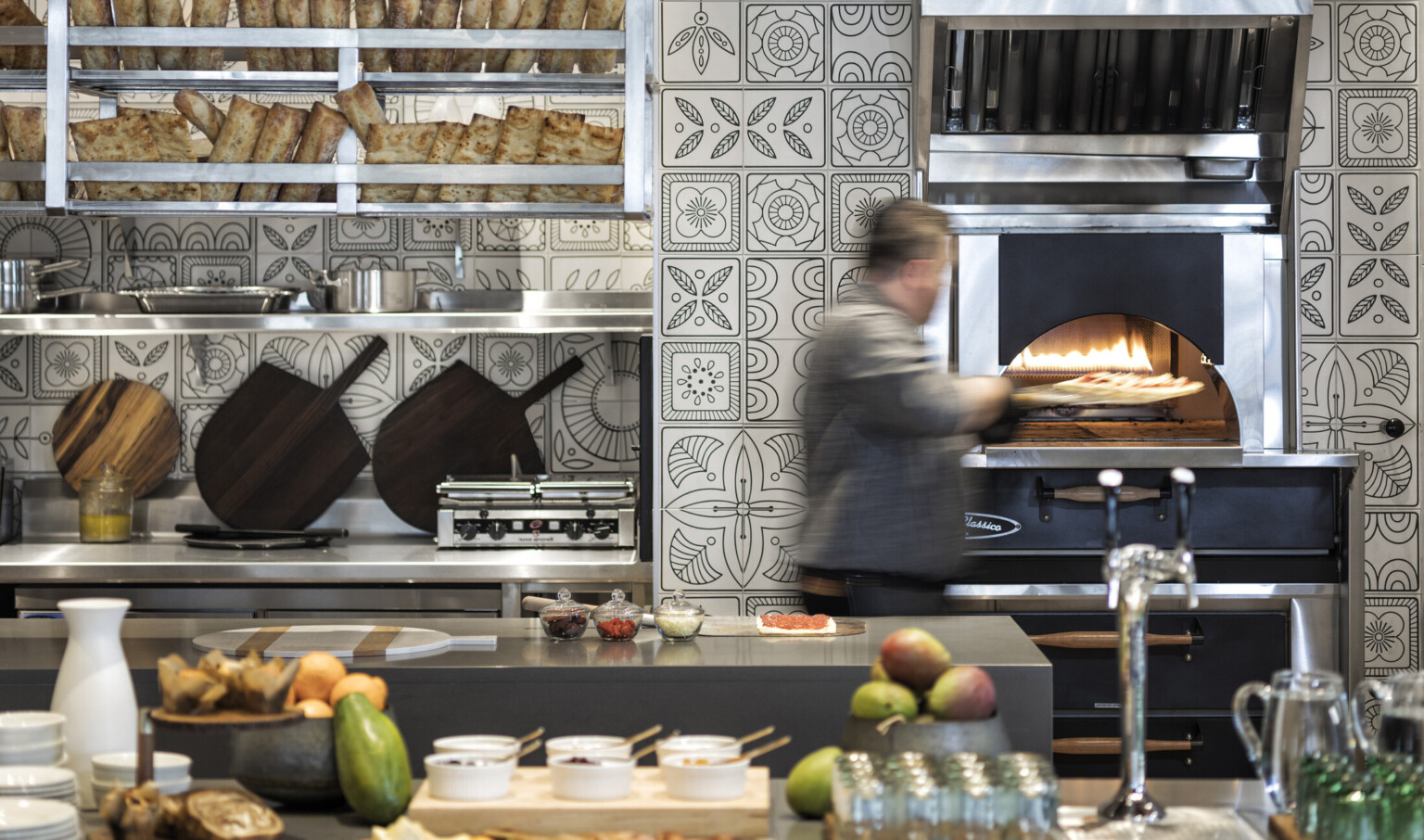 Pizza being placed into oven in kitchen with black and white floral tiles. Stainless steel counters and shelving with bread