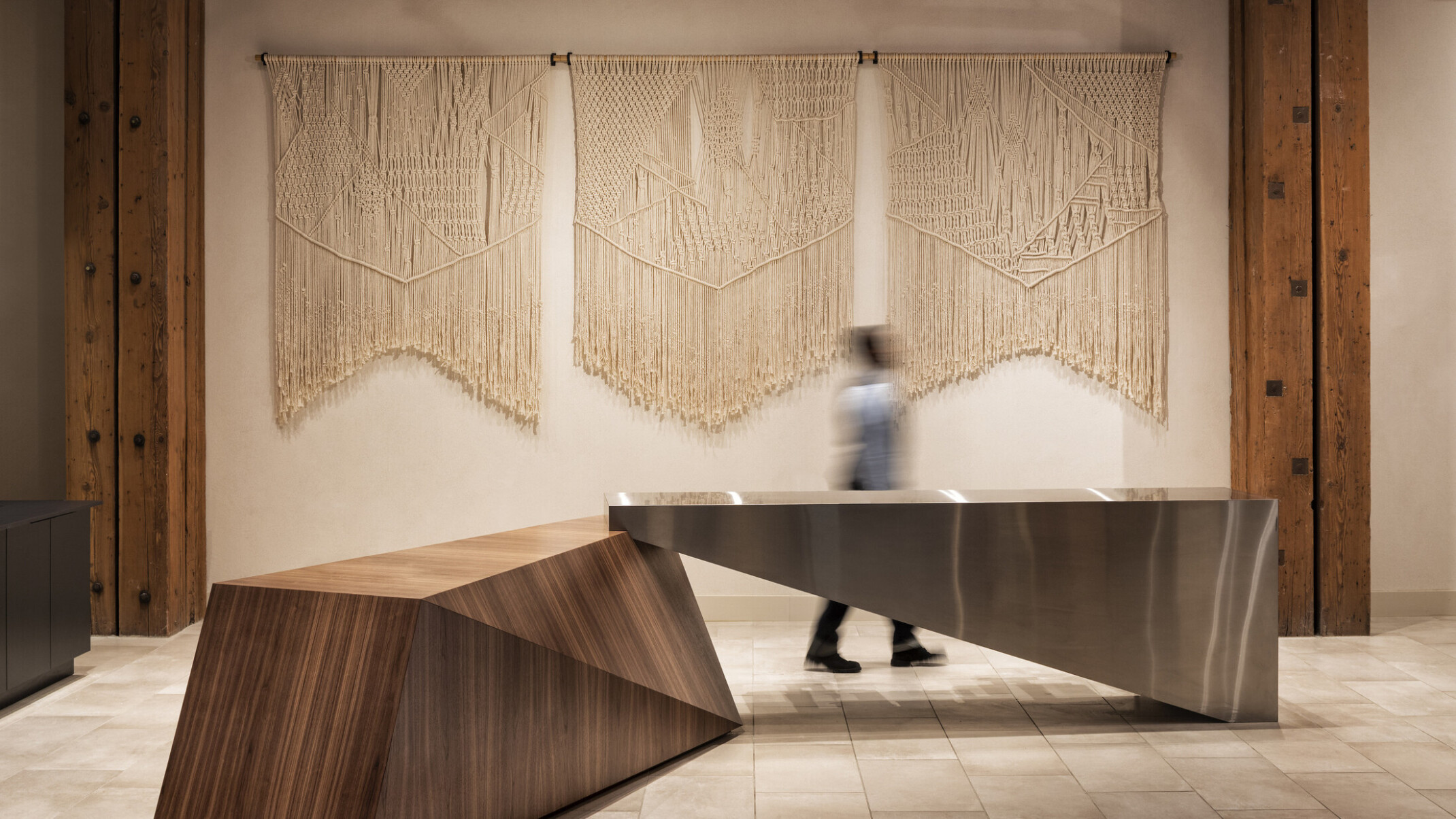 White wall with macrame wall hanging between wood pillars on tile floor with geometric wood and metal table at center