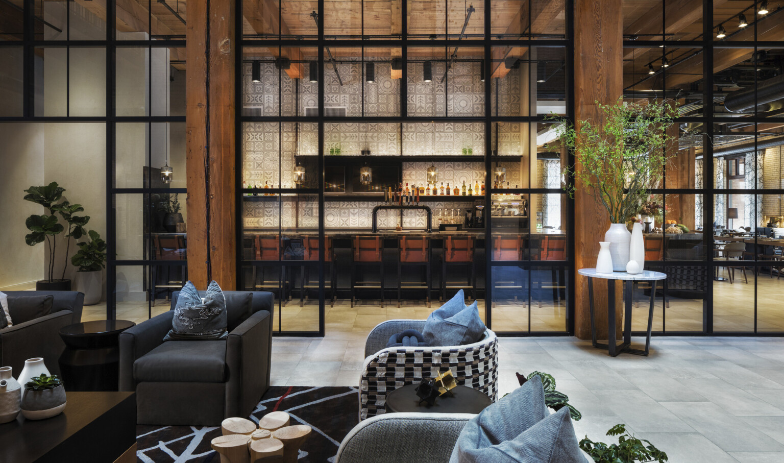 Seating area in room with double height windows looking to white geometric walled bar in next room. Wood columns and ceiling
