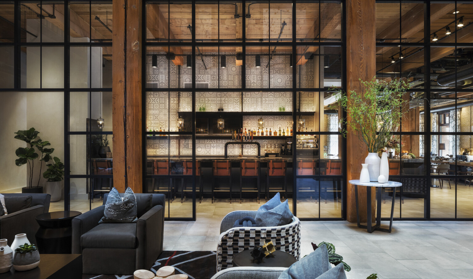 Seating area in room with double height windows looking to white geometric walled bar in next room. Wood columns and ceiling