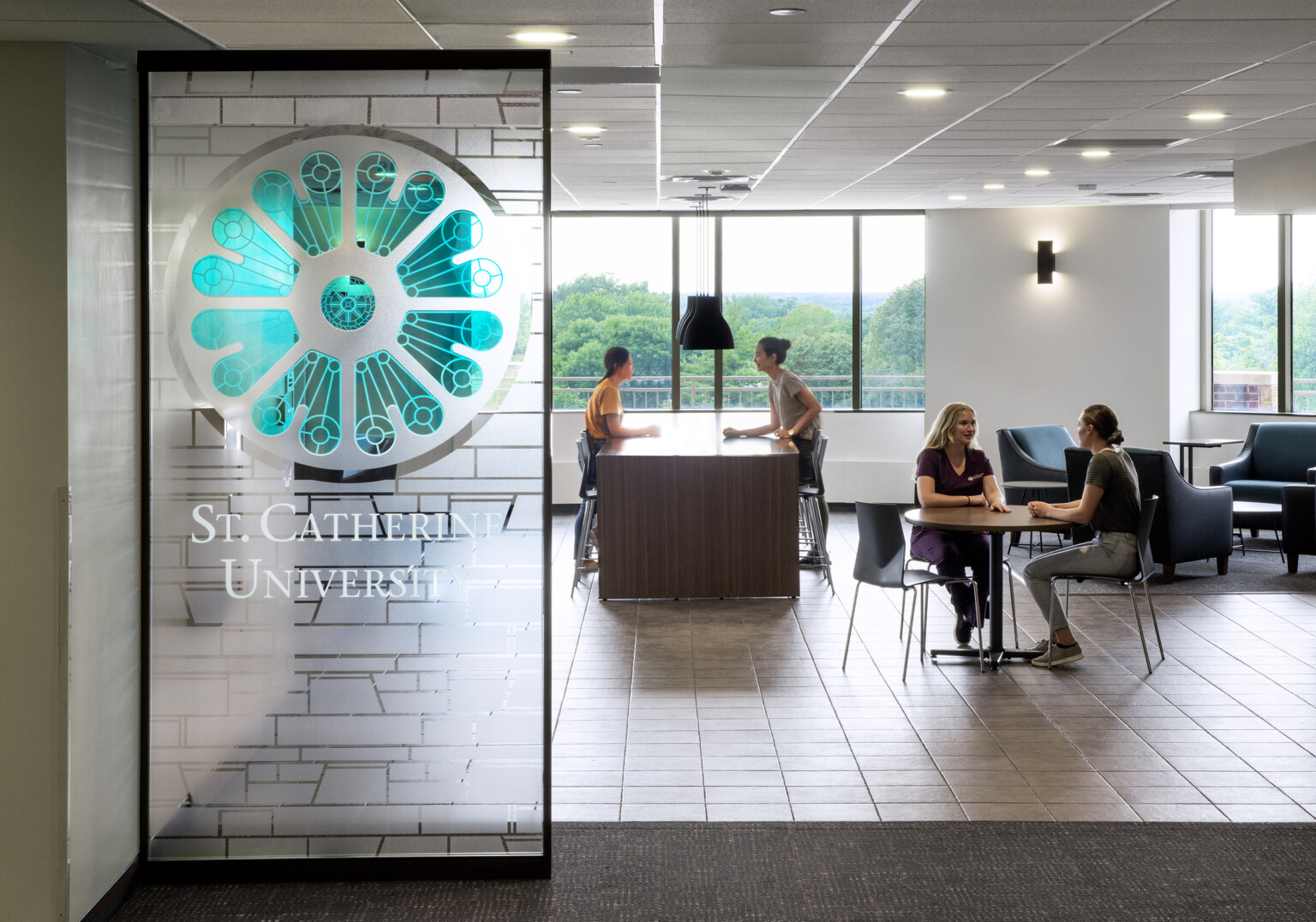Entrance to a common space of St. Catherine University; students sitting at tables with windows looking out to nature; frosted white and green glass entrance sign