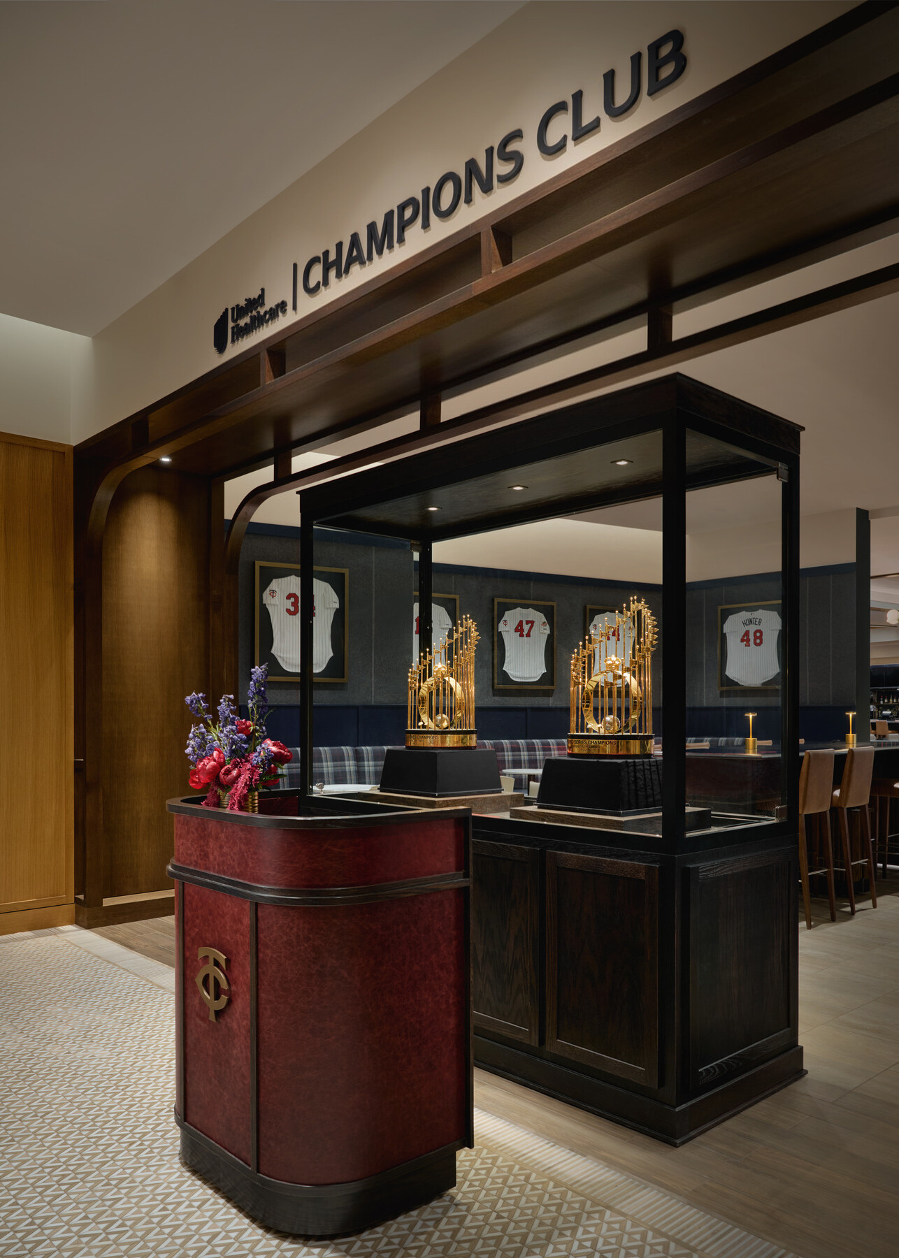 Entrance to the UnitedHealthcare Champions Club featuring a trophy display case, Minnesota Twins branding, framed jerseys, and upscale wood and leather finishes.