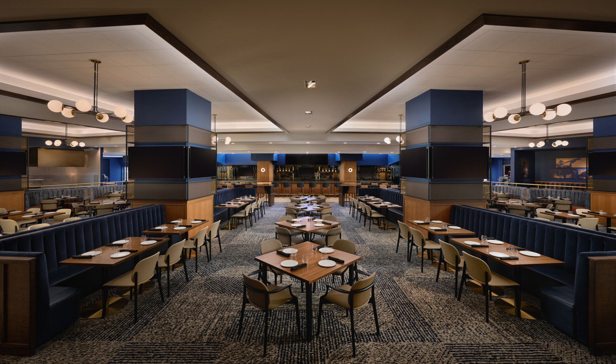 Spacious upscale dining lounge with booth and table seating, large display screens, blue accent columns, and modern globe pendant lighting.