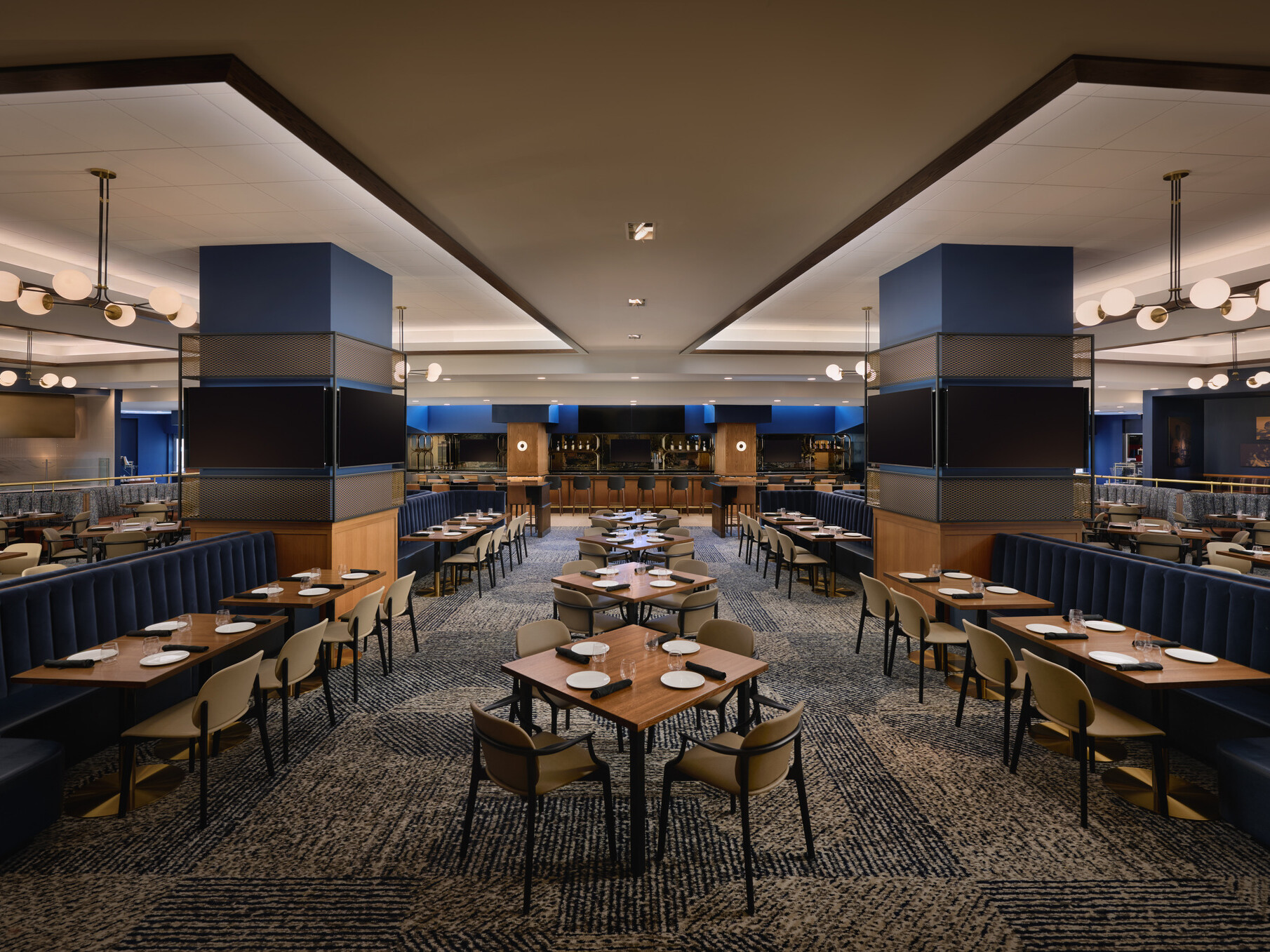 Spacious upscale dining lounge with booth and table seating, large display screens, blue accent columns, and modern globe pendant lighting.