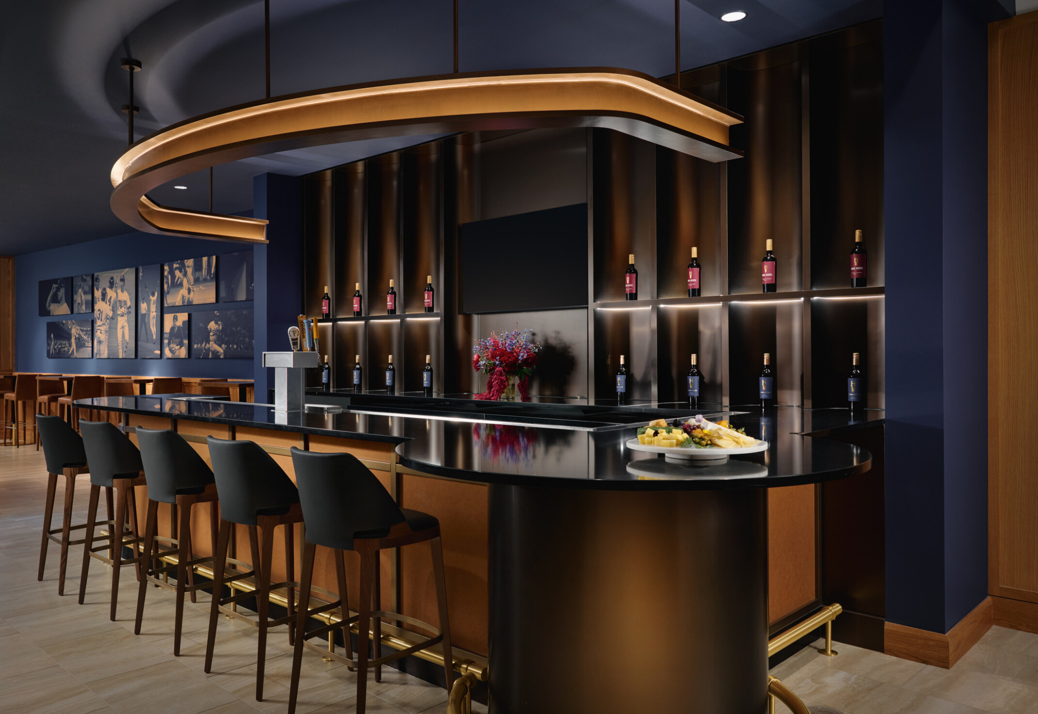Elegant hospitality bar with five stools, illuminated wine bottle displays, beverage taps, a large decorative overhead light, and a glossy black counter featuring a cheese plate and flowers. The space combines dark blue walls, warm wood details, and ambient lighting for a refined tasting-room setting.