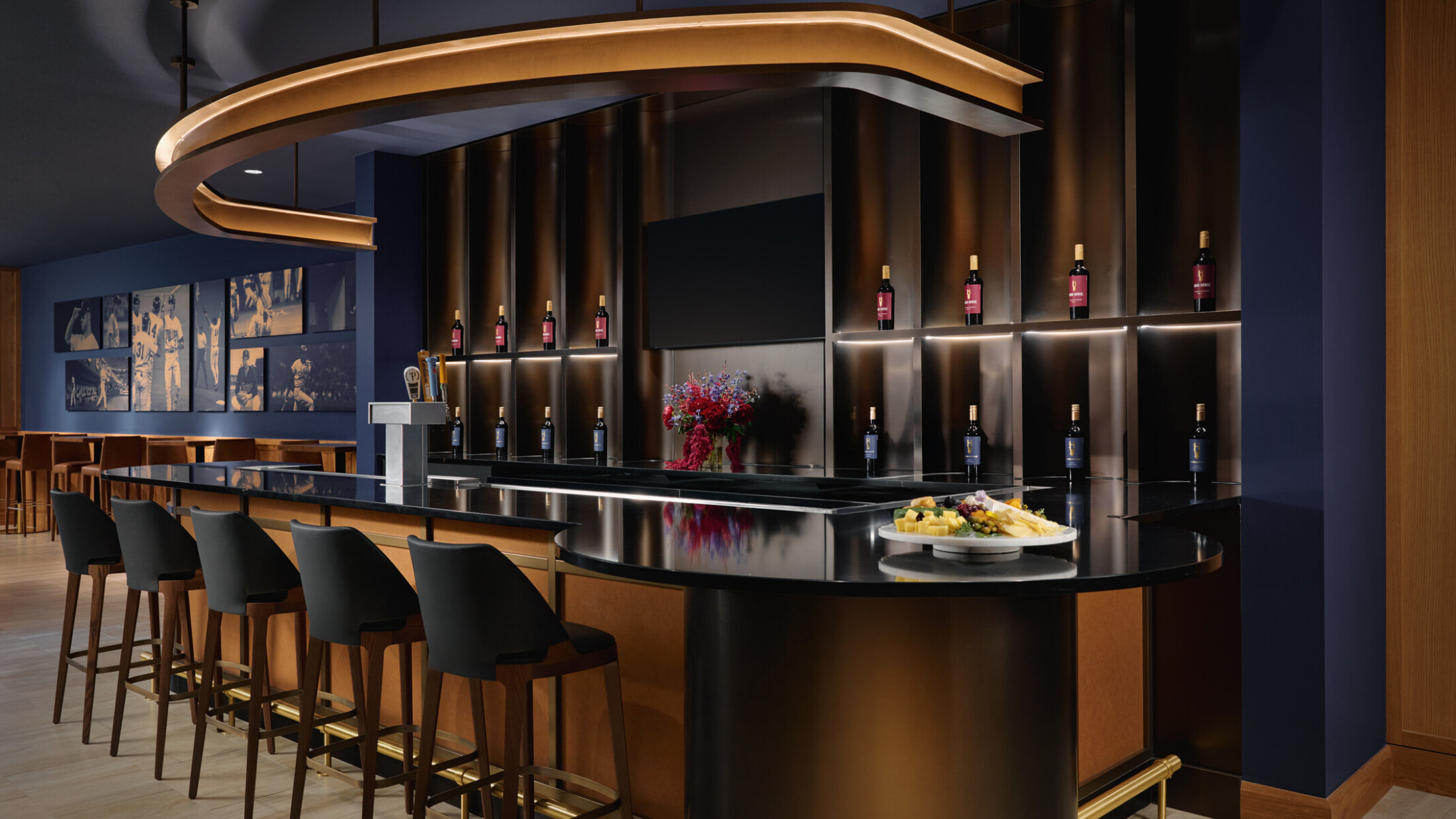Elegant hospitality bar with five stools, illuminated wine bottle displays, beverage taps, a large decorative overhead light, and a glossy black counter featuring a cheese plate and flowers. The space combines dark blue walls, warm wood details, and ambient lighting for a refined tasting-room setting.