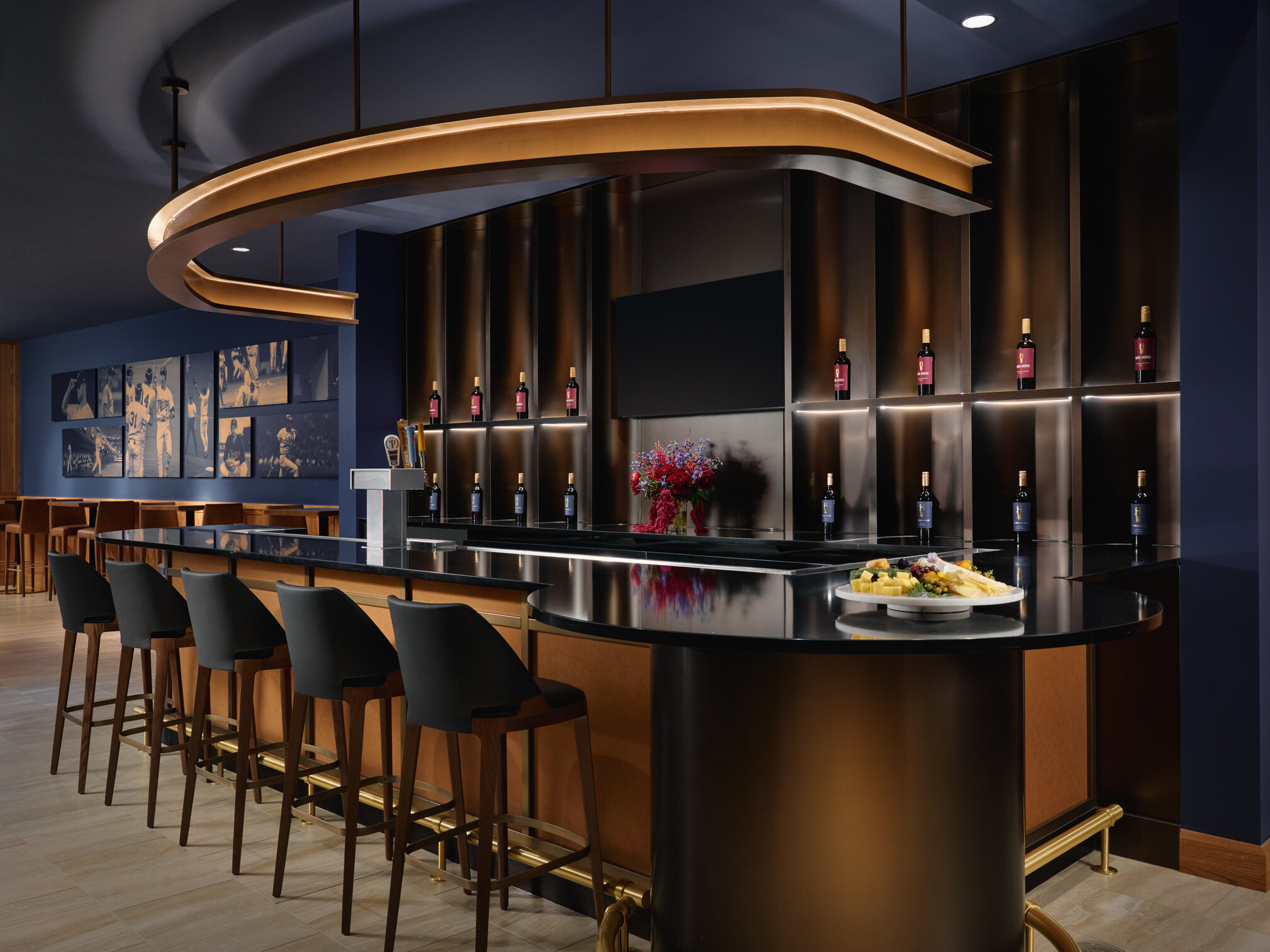 Elegant hospitality bar with five stools, illuminated wine bottle displays, beverage taps, a large decorative overhead light, and a glossy black counter featuring a cheese plate and flowers. The space combines dark blue walls, warm wood details, and ambient lighting for a refined tasting-room setting.