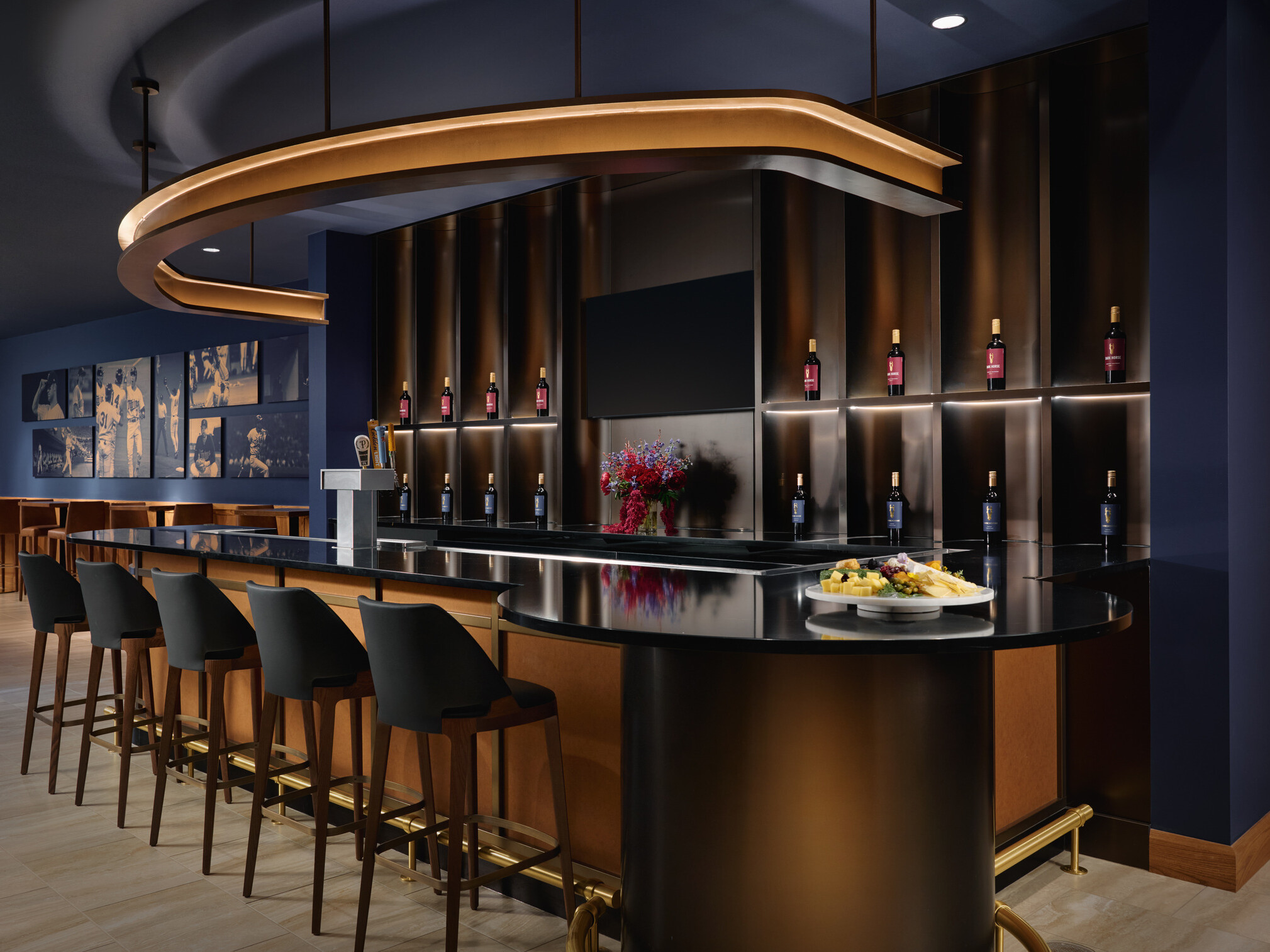 Elegant hospitality bar with five stools, illuminated wine bottle displays, beverage taps, a large decorative overhead light, and a glossy black counter featuring a cheese plate and flowers. The space combines dark blue walls, warm wood details, and ambient lighting for a refined tasting-room setting.