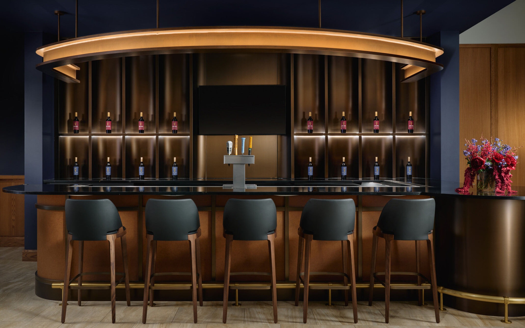 Modern tasting bar with five dark barstools facing a glossy black counter. Backlit shelving displays wine bottles on both sides of a central television screen and beer taps, beneath a large circular pendant light. Dark blue walls, warm wood finishes, and a floral arrangement create an upscale atmosphere.