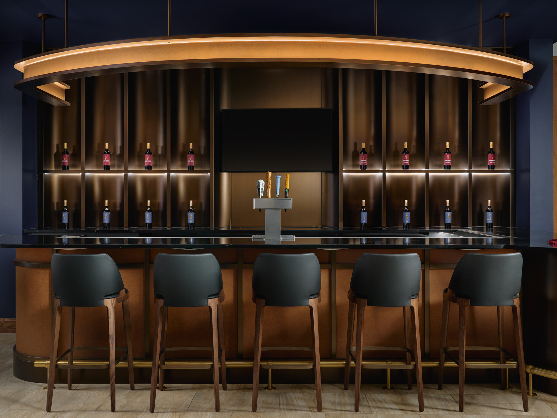 Modern tasting bar with five dark barstools facing a glossy black counter. Backlit shelving displays wine bottles on both sides of a central television screen and beer taps, beneath a large circular pendant light. Dark blue walls, warm wood finishes, and a floral arrangement create an upscale atmosphere.