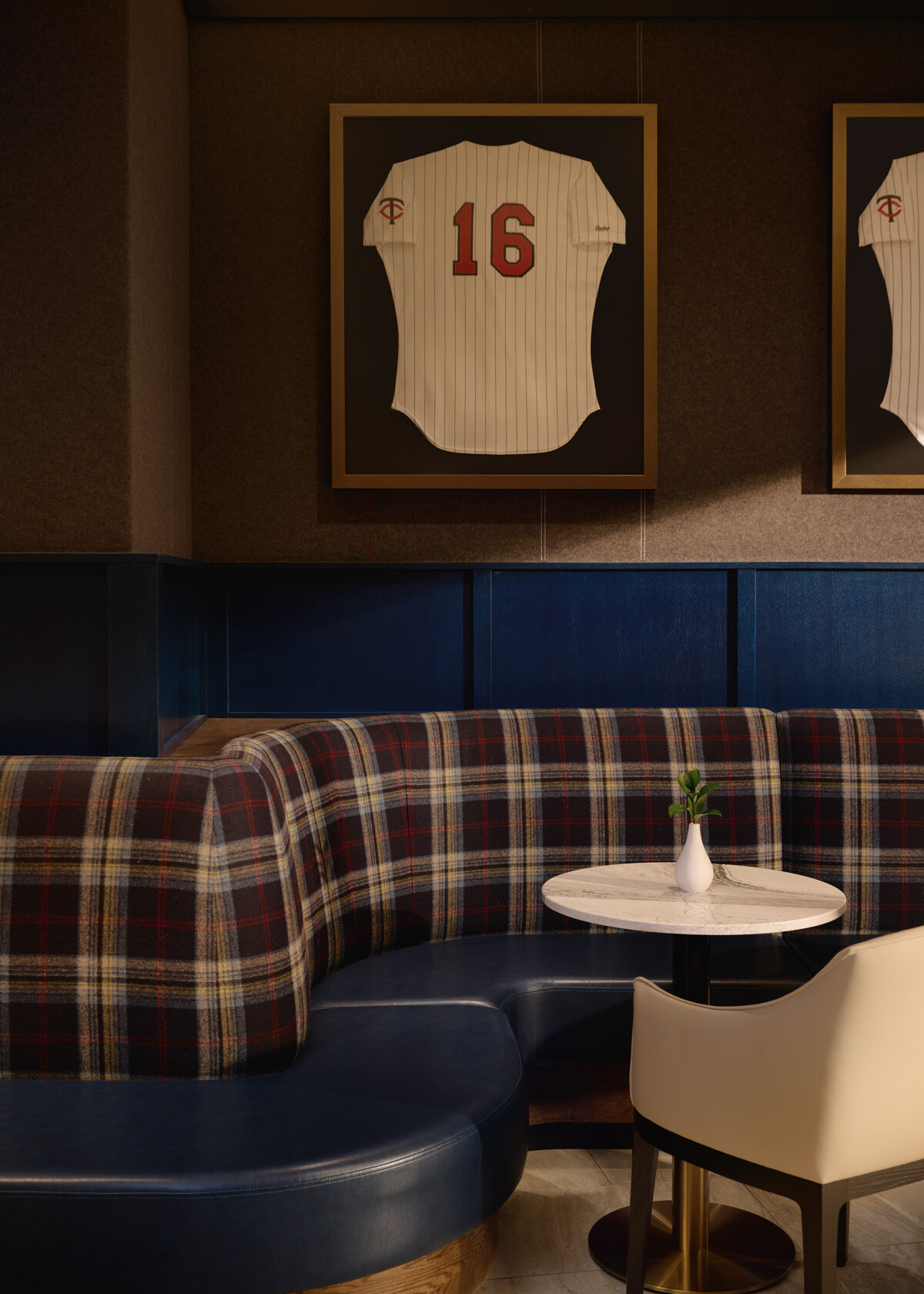 Curved booth seating with blue leather cushions and plaid upholstery surrounds a small marble table beneath framed Minnesota Twins jersey displays in a refined lounge setting.