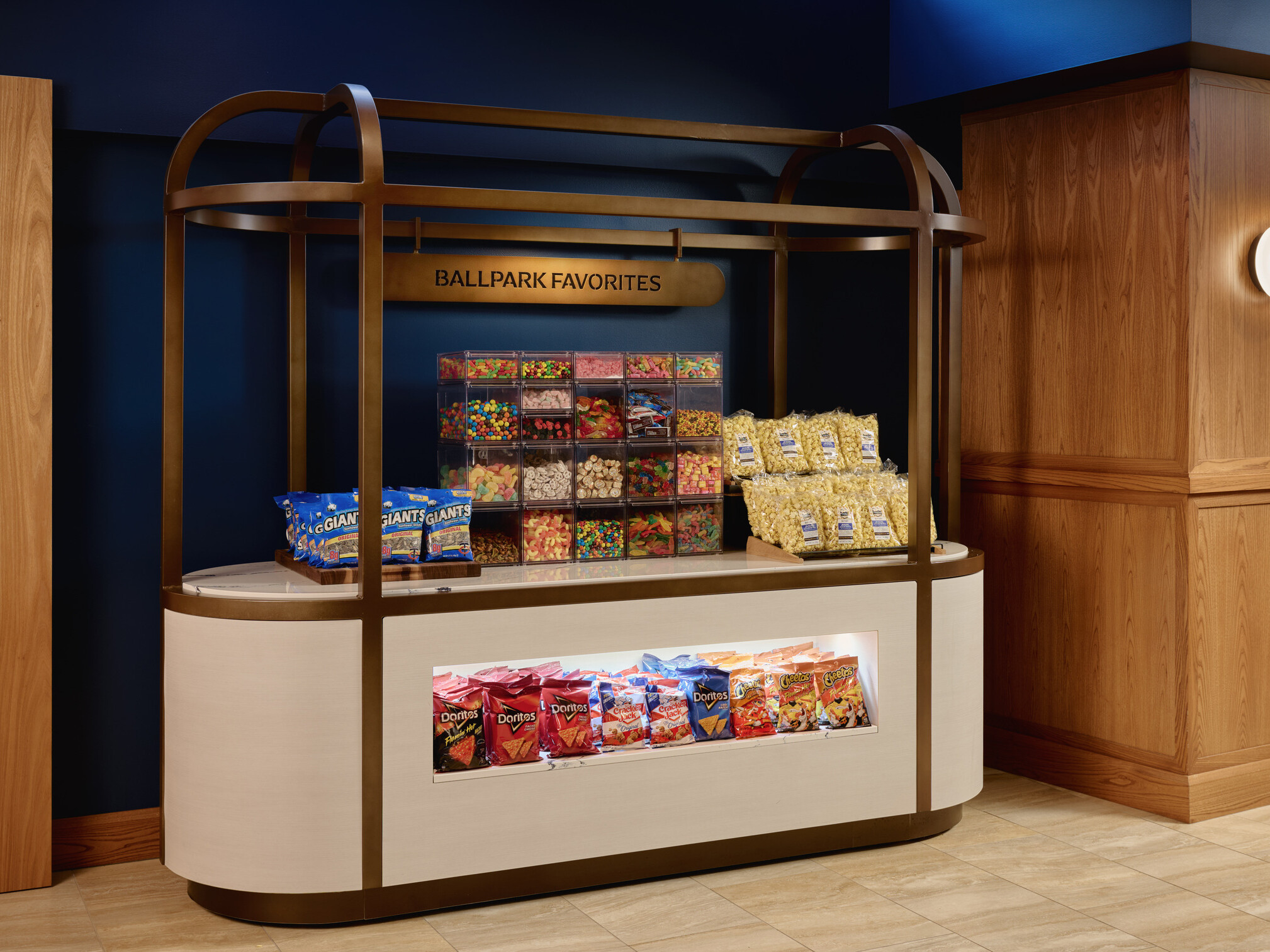 Snack station labeled “Ballpark Favorites” featuring packaged popcorn, candy bins, and assorted chips within a modern concession-style display against dark blue walls and wood paneling.