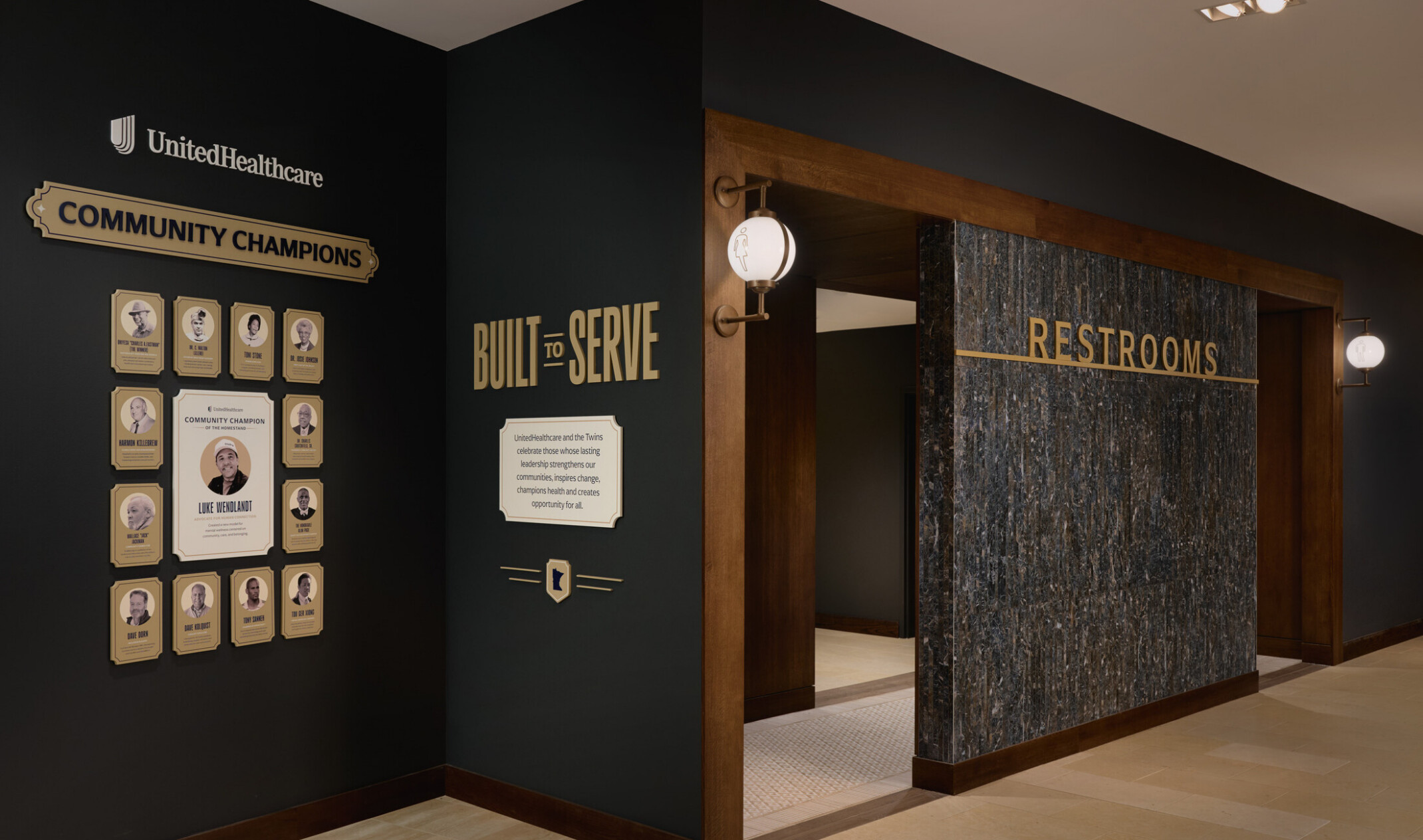 Hallway entrance to restrooms featuring wood and stone finishes, with recognition displays for UnitedHealthcare Community Champions and a “Built to Serve” community program.