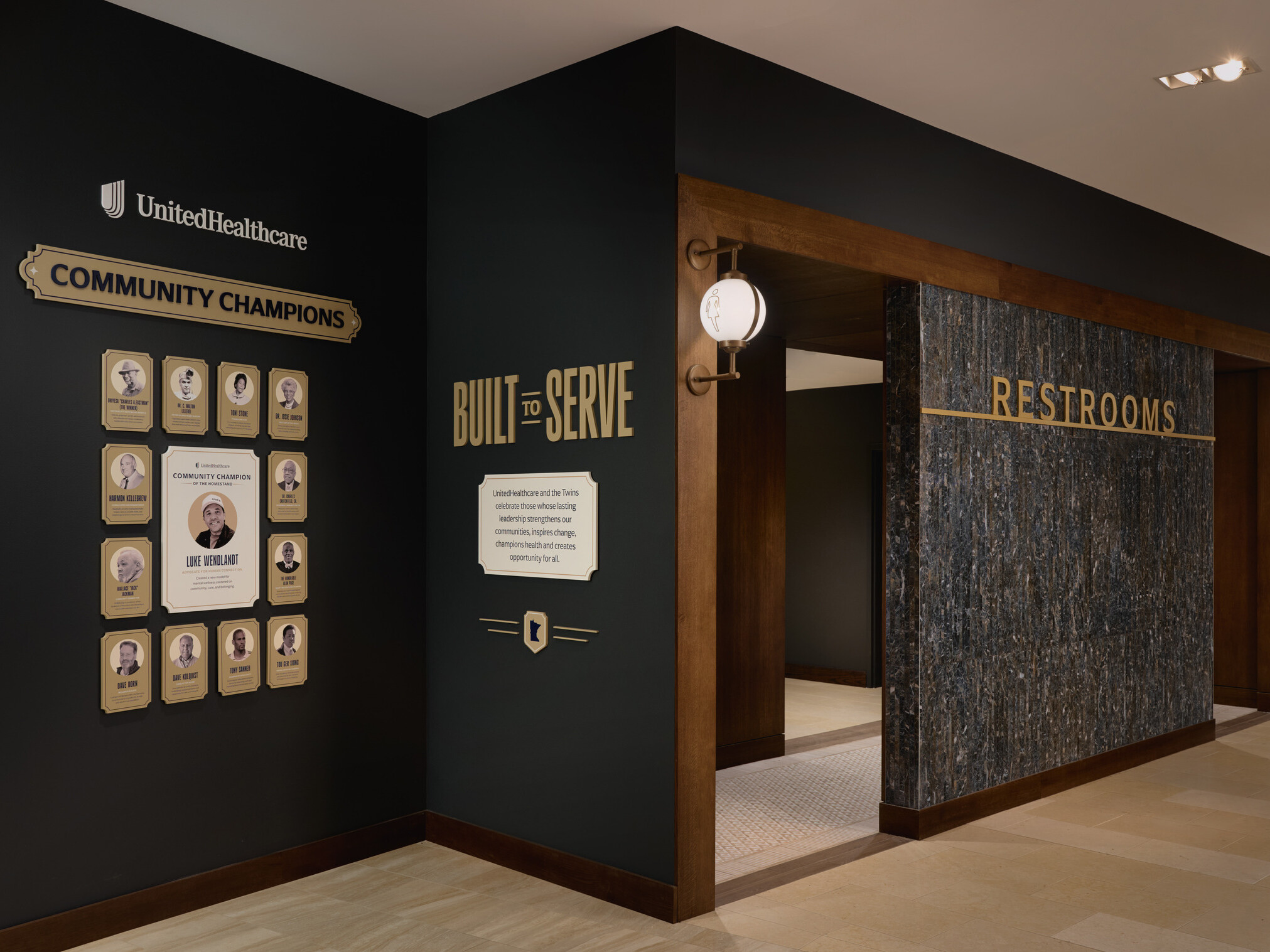 Hallway entrance to restrooms featuring wood and stone finishes, with recognition displays for UnitedHealthcare Community Champions and a “Built to Serve” community program.