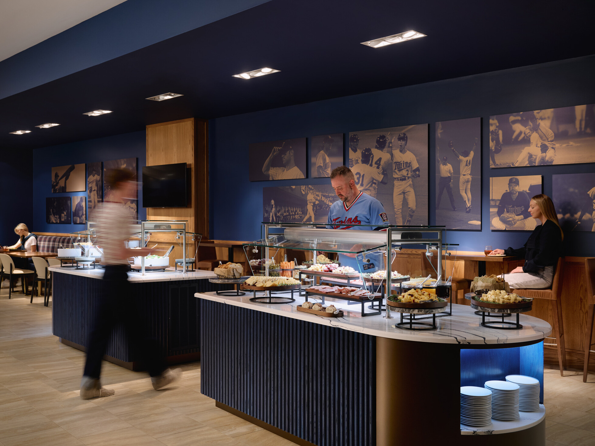 Modern buffet lounge with glass-covered food stations, guests dining and serving themselves, and large baseball-themed wall art displayed against dark blue walls.
