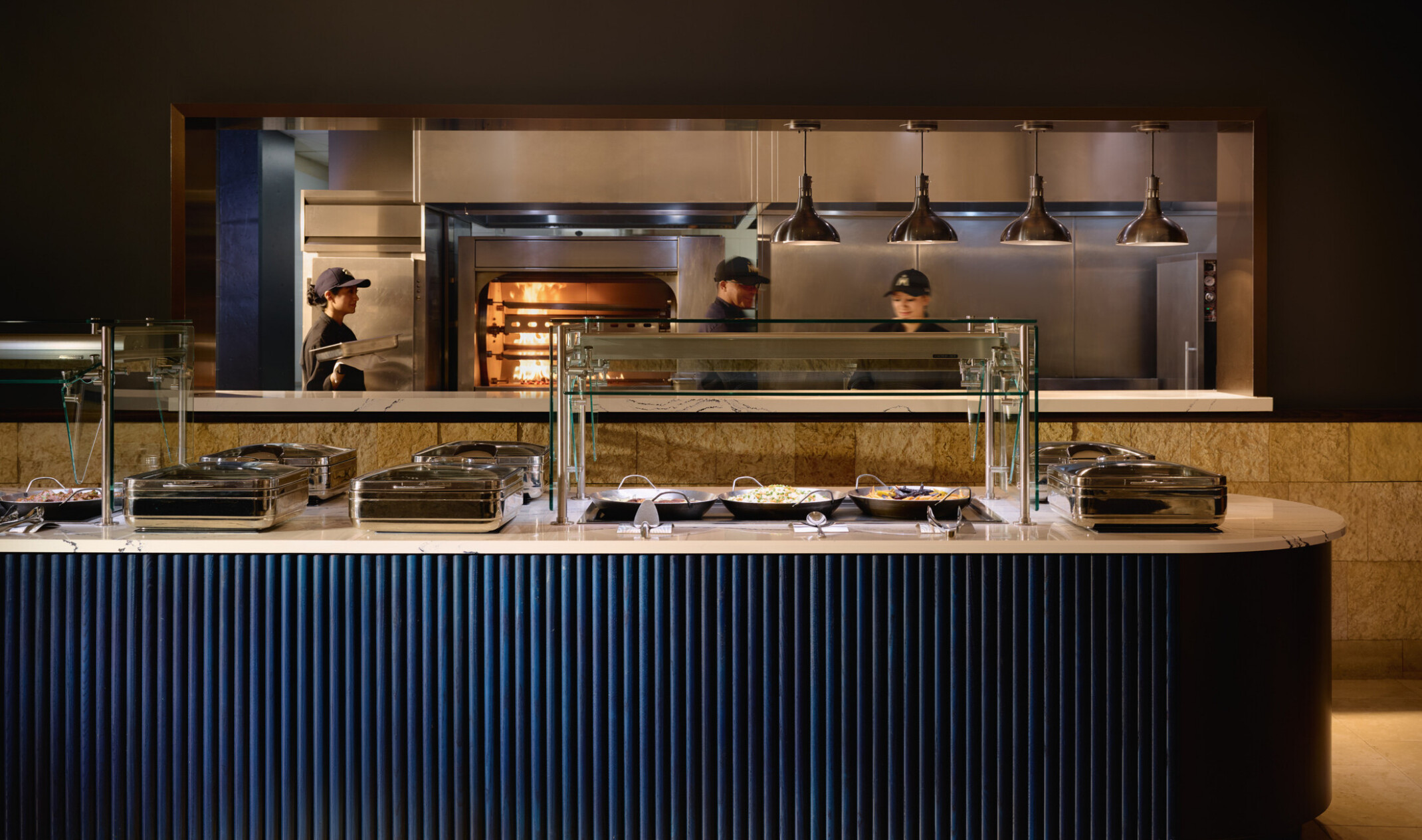 Modern buffet station with glass food guards and stainless-steel serving trays in front of an open kitchen. Three staff members work near a wood-fired oven beneath industrial pendant lights.