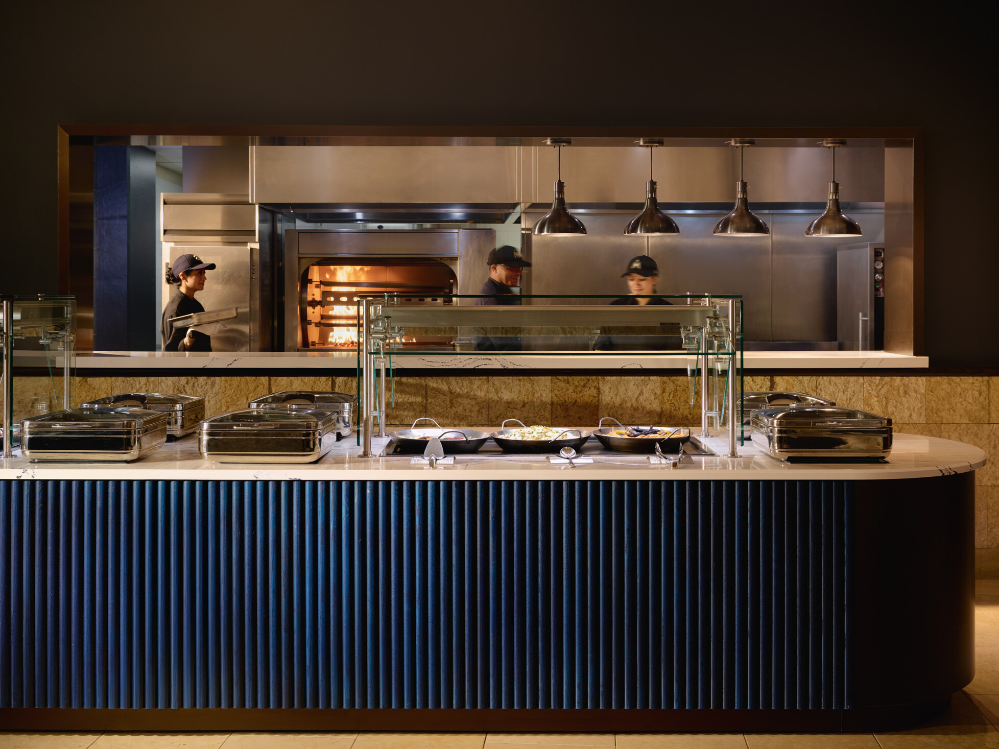 Modern buffet station with glass food guards and stainless-steel serving trays in front of an open kitchen. Three staff members work near a wood-fired oven beneath industrial pendant lights.