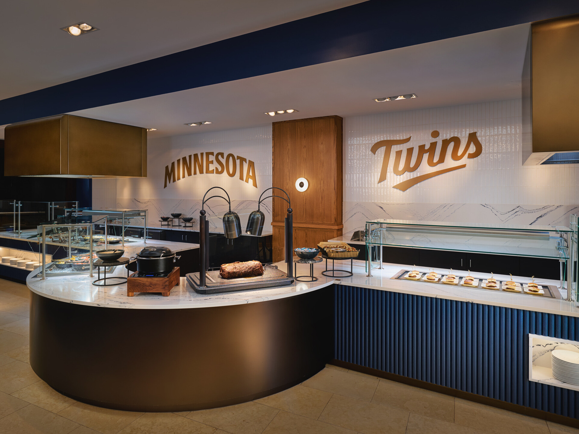 Contemporary buffet area with curved serving counters, glass food displays, and carved wall signage reading “Minnesota” and “Twins.” A carved roast and plated appetizers are displayed under warming lamps in a modern hospitality setting.