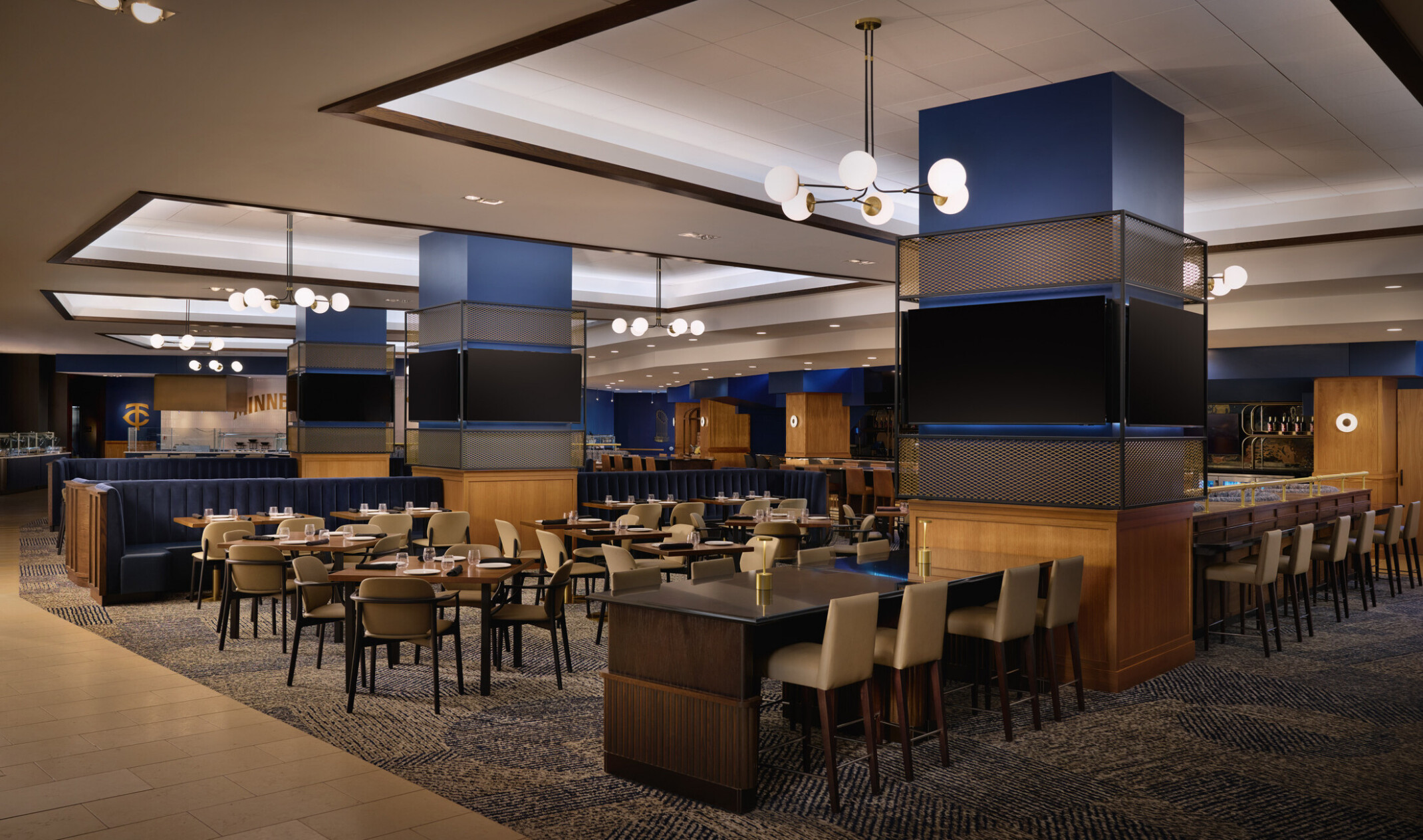 Spacious upscale dining lounge with booth and table seating, large display screens, blue accent columns, and modern globe pendant lighting.