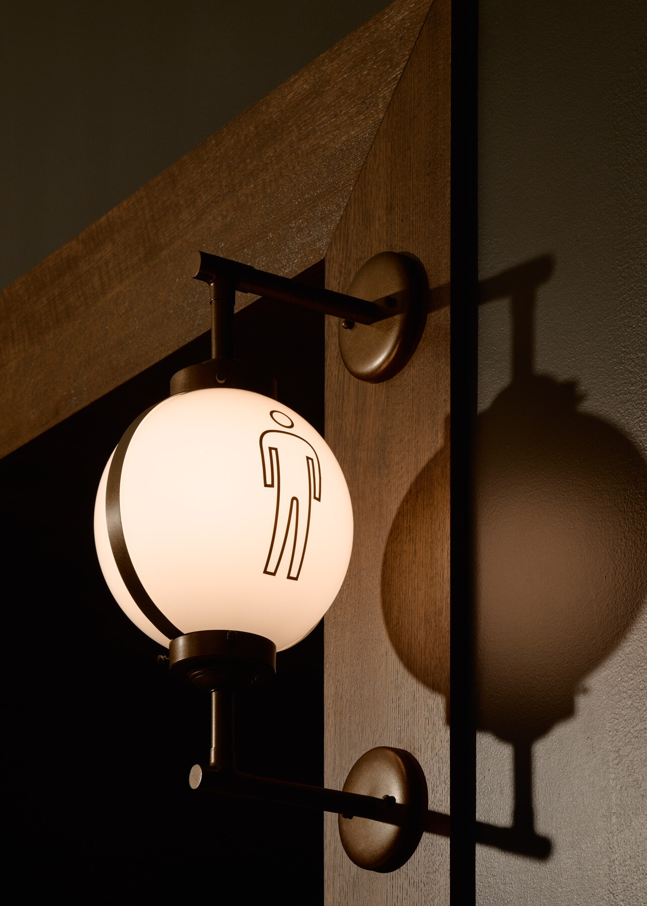 Decorative illuminated wall sconce featuring a restroom accessibility symbol, mounted on a dark wall with warm wood trim and casting a shadow across the surface.
