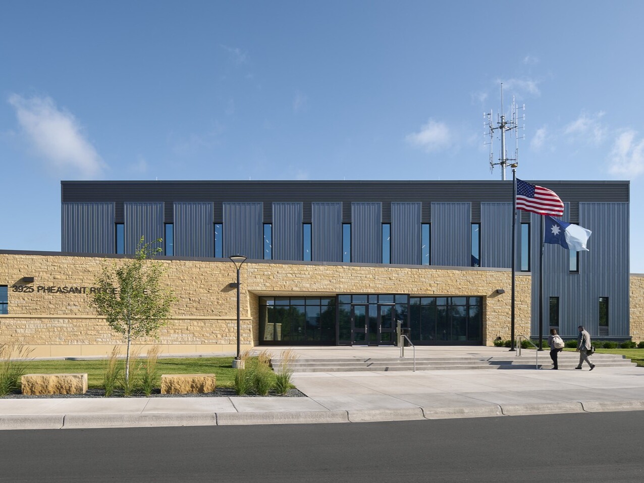 Bright blue with white clouds above a large building made of mixed materials; 2 people walk toward the entrance stairs, where two flagpoles stand in front of the doors; landscaping around the building is kept
