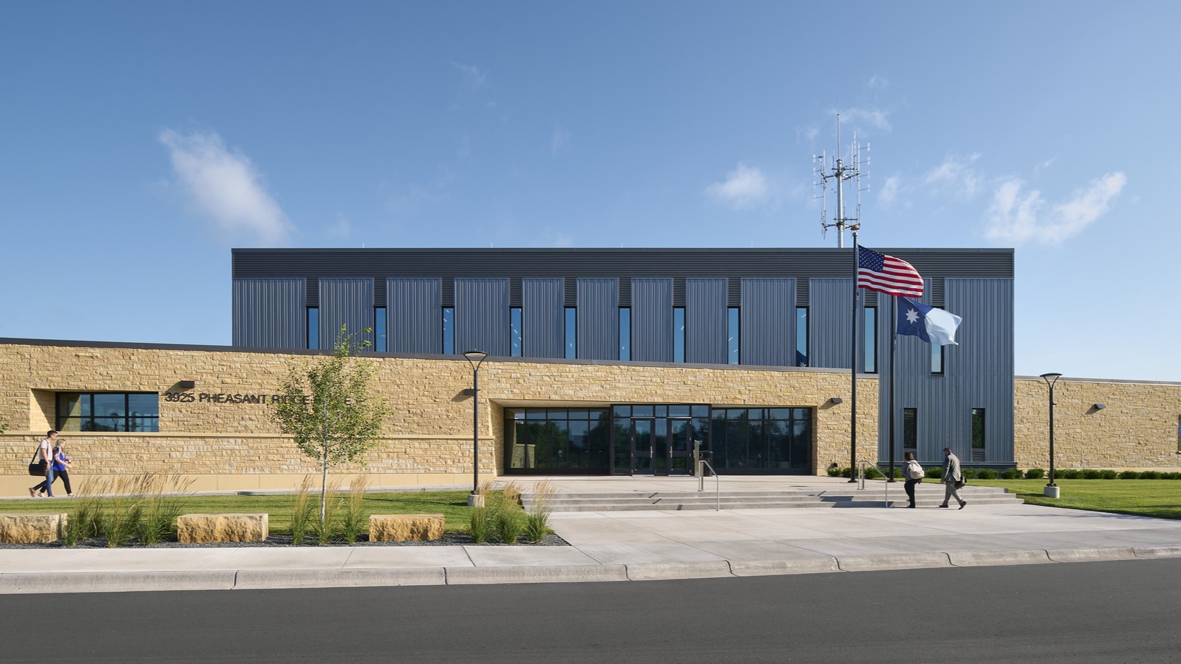 Bright blue with white clouds above a large building made of mixed materials; 2 people walk toward the entrance stairs, where two flagpoles stand in front of the doors; landscaping around the building is kept