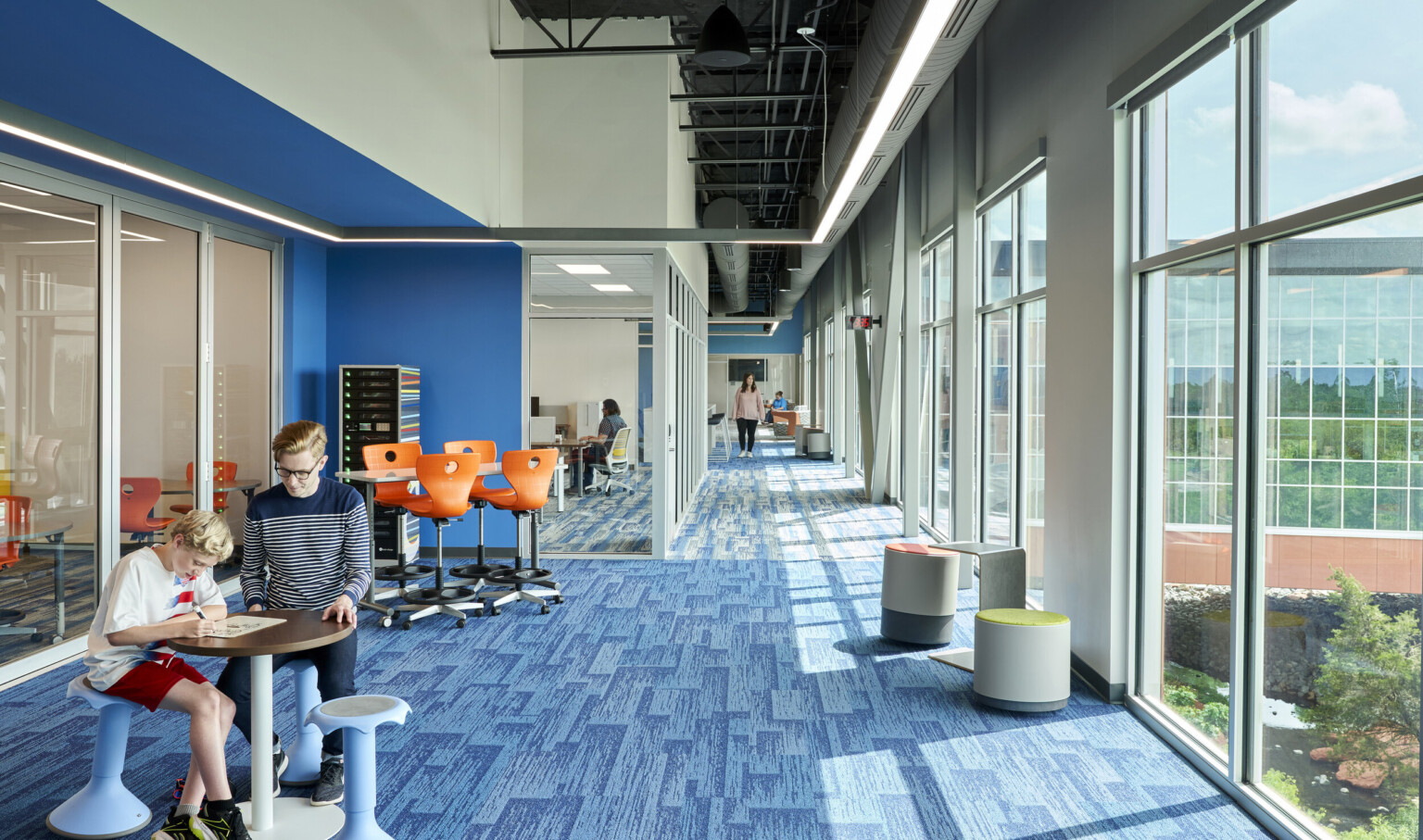 Blue carpeted hall with large windows on right wall. Left, recessed blue wall accent along glass wall looking into classes