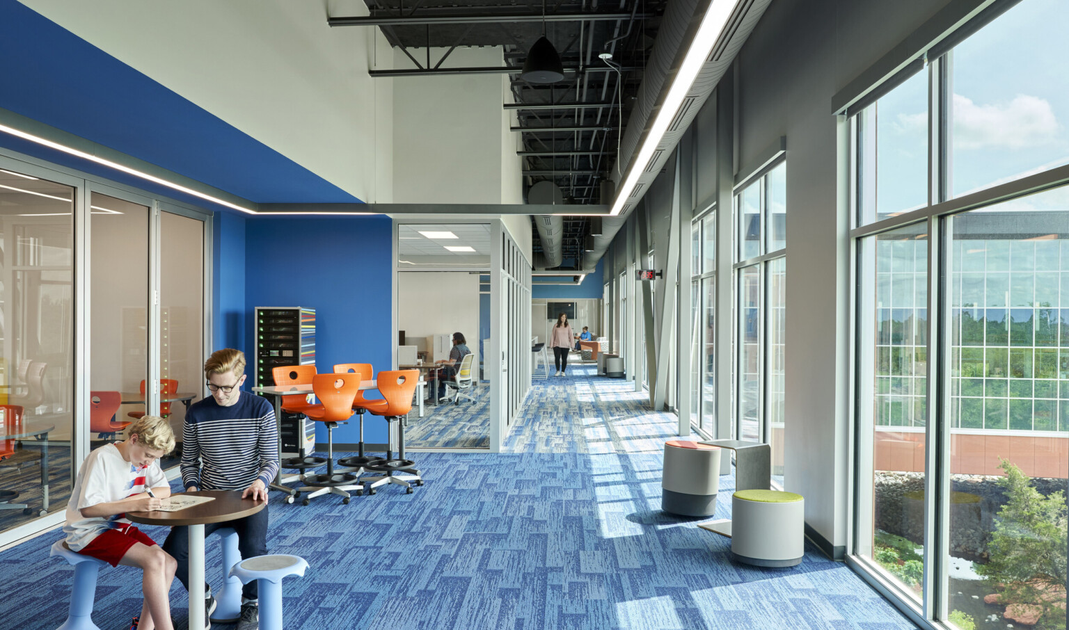 Blue carpeted hall with large windows on right wall. Left, recessed blue wall accent along glass wall looking into classes Blue carpeted hall with large windows on right wall. Left, recessed blue wall accent along glass wall looking into classes