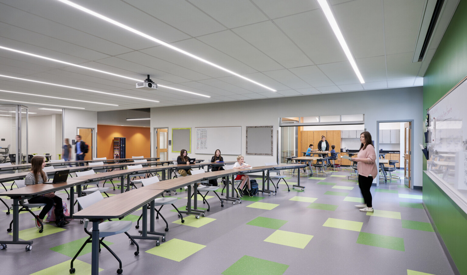Classroom with green accent wall and floor tiles. Orange walls in hallway through flexible walls. Rolling chairs and long tables