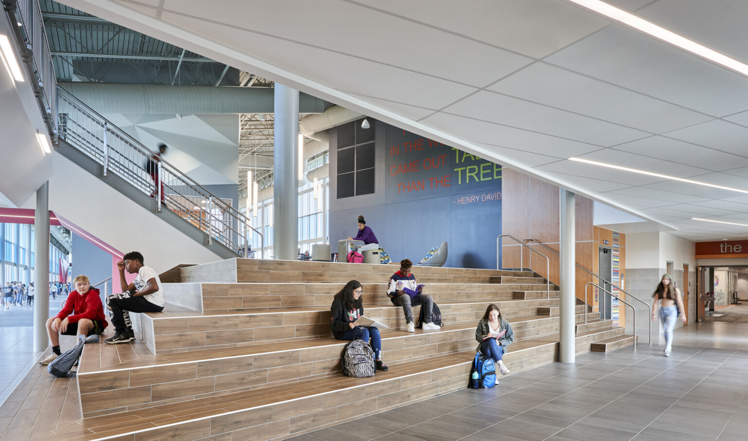 Students sit on indoor learning stair between leading to muraled wall in double height lobby with mezzanine