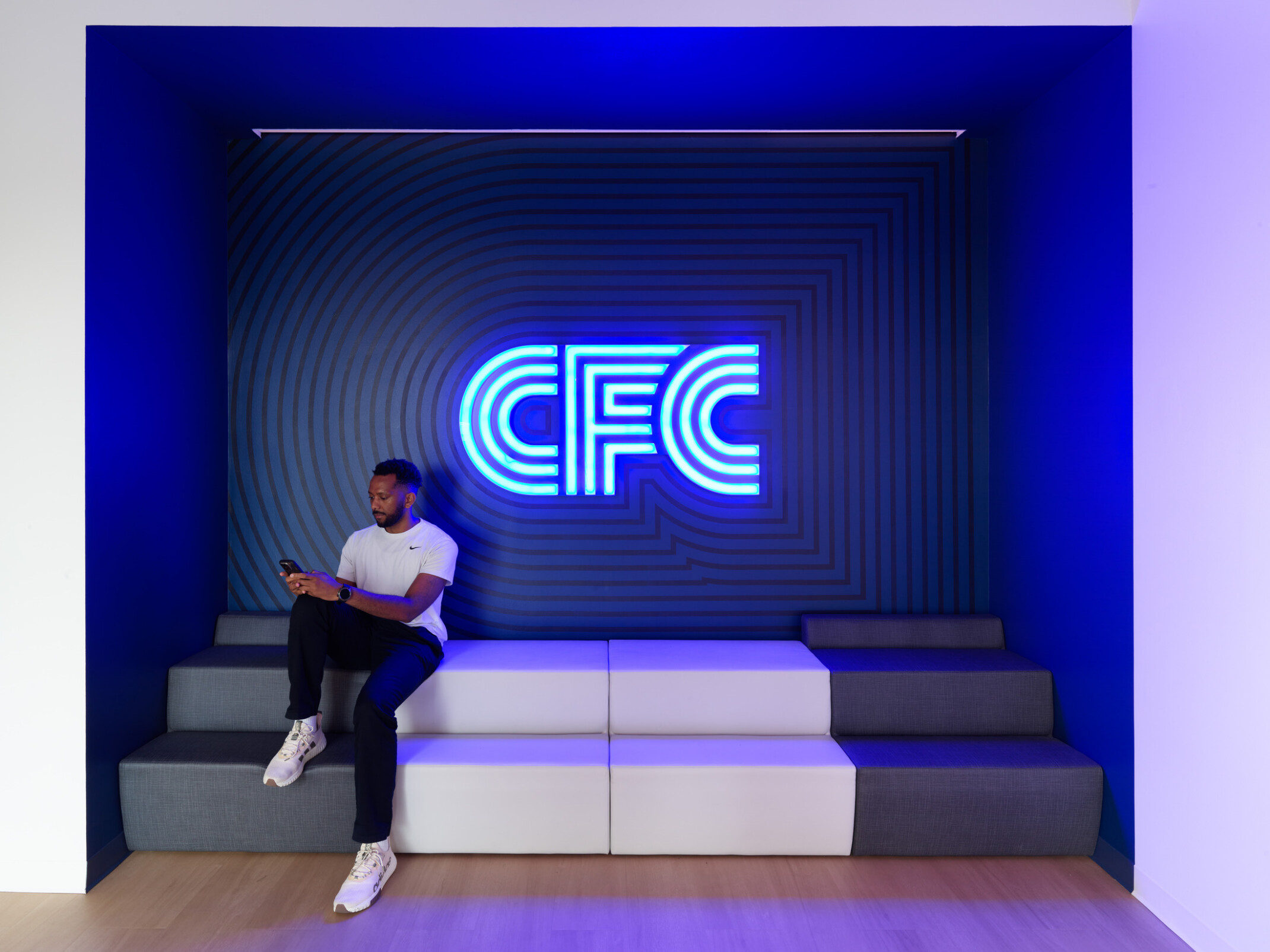Built-in seating alcove featuring a glowing blue “CFC” sign, geometric wall pattern, and tiered bench seating, with a person seated and looking at a mobile phone