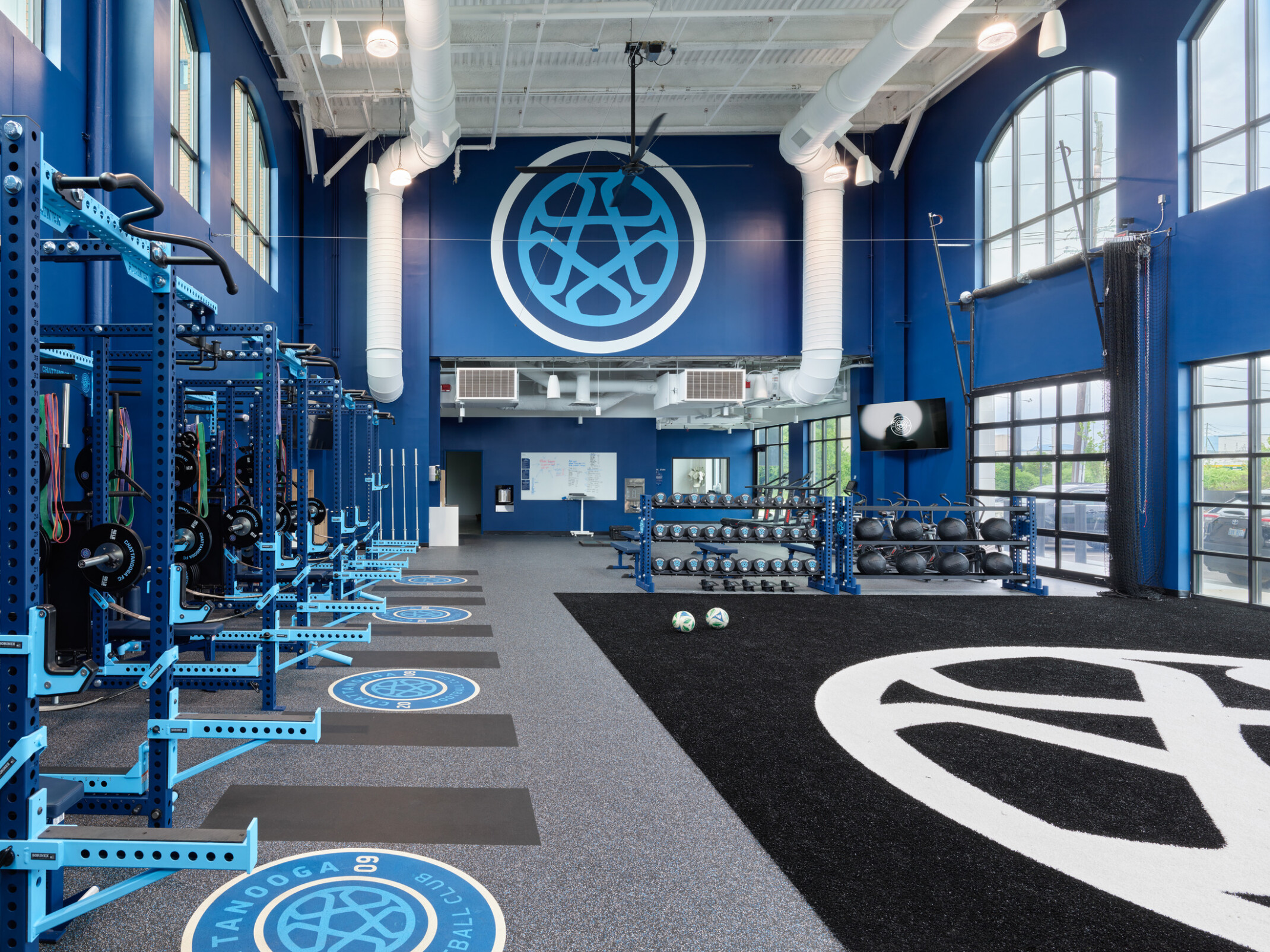 Large Chattanooga Football Club training facility featuring blue-branded strength equipment, free weights, turf training space, and a prominent club logo displayed on a tall accent wall.