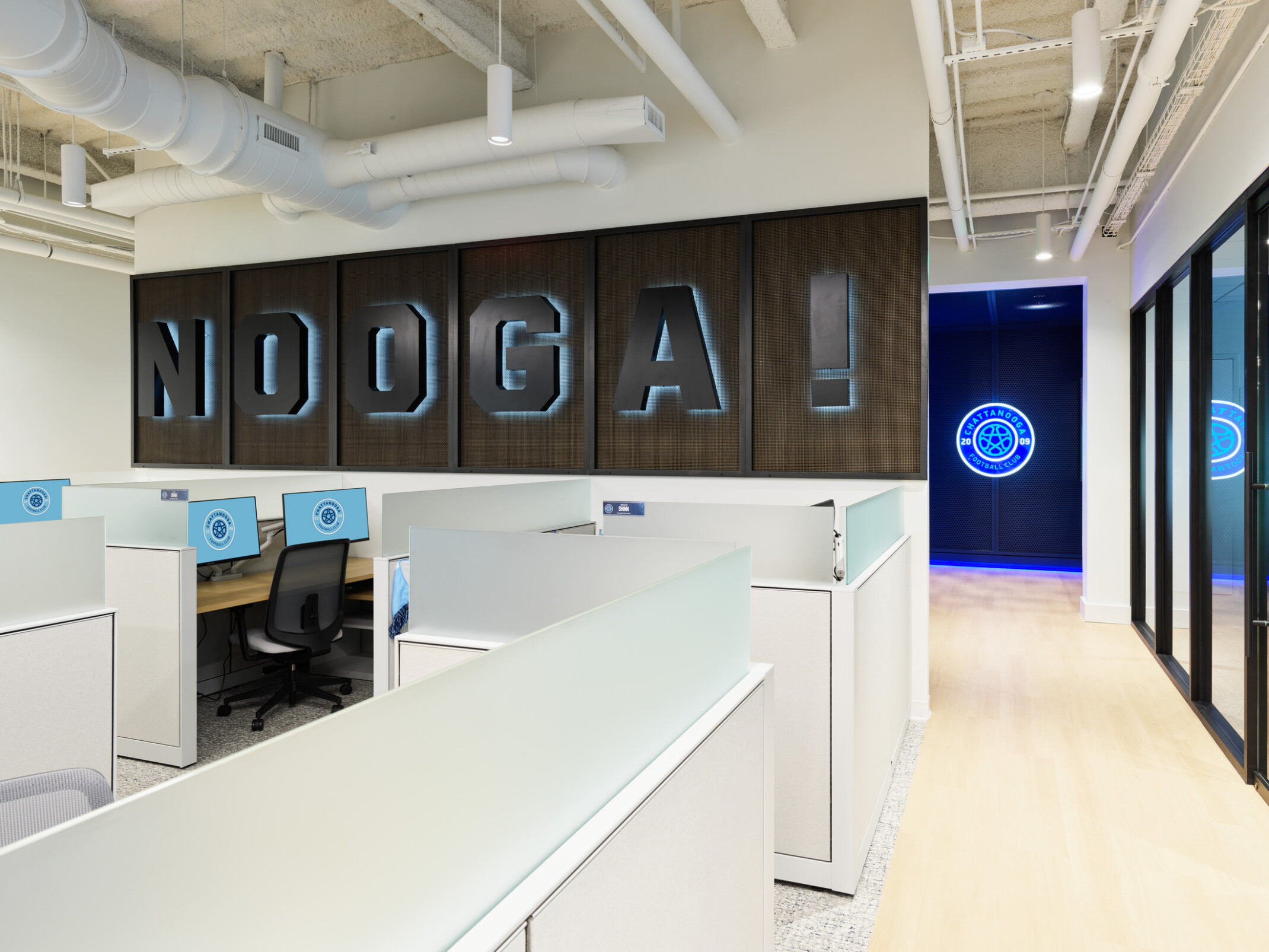 Open office workspace with cubicles beneath a large illuminated wall sign reading “NOOGA!”, featuring Chattanooga Football Club branding and a glowing club logo visible down the hallway