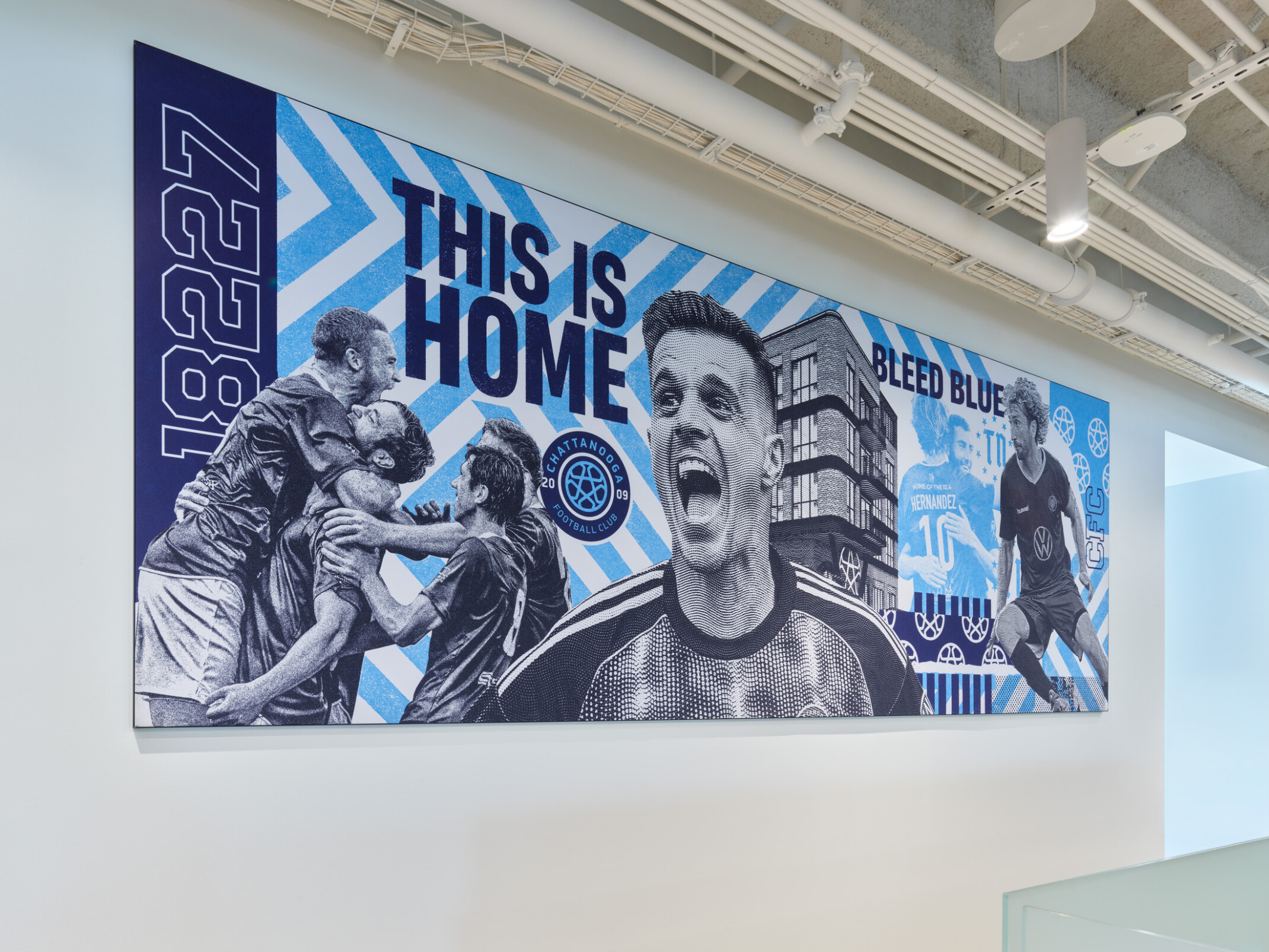 Large Chattanooga Football Club mural featuring action soccer imagery, club branding, and the slogans “THIS IS HOME” and “BLEED BLUE” displayed on an office wall.