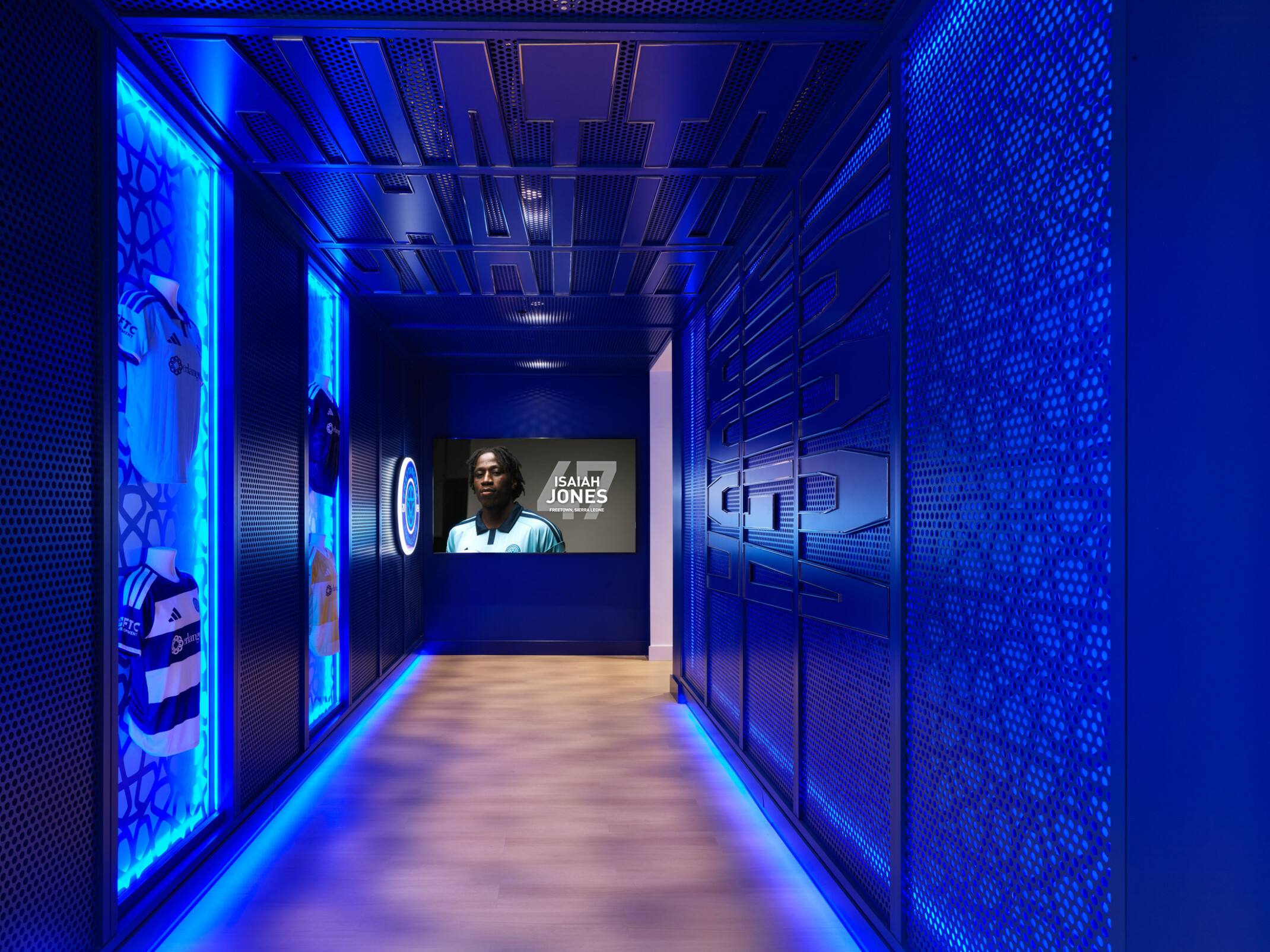 Blue-lit hallway in a Chattanooga Football Club facility featuring illuminated display cases with team jerseys, perforated metal wall panels, and a digital screen showing a player profile at the end of the corridor.
