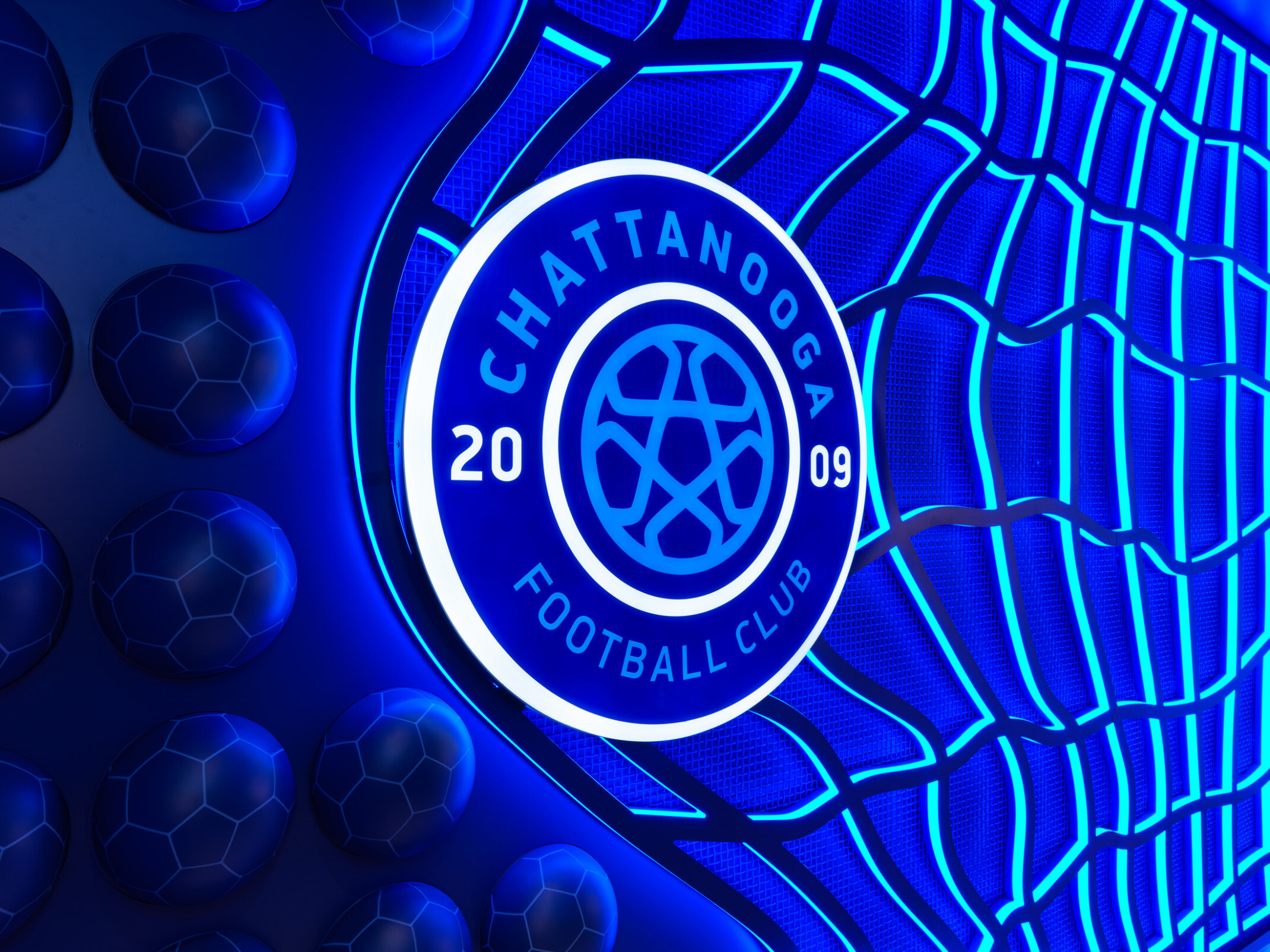 Illuminated blue logo for Chattanooga Football Club mounted against a decorative background featuring a stylized soccer net and raised soccer-ball patterns. The circular emblem displays the text “CHATTANOOGA FOOTBALL CLUB” and “2009” around a soccer ball design, glowing with bright blue and white lighting.