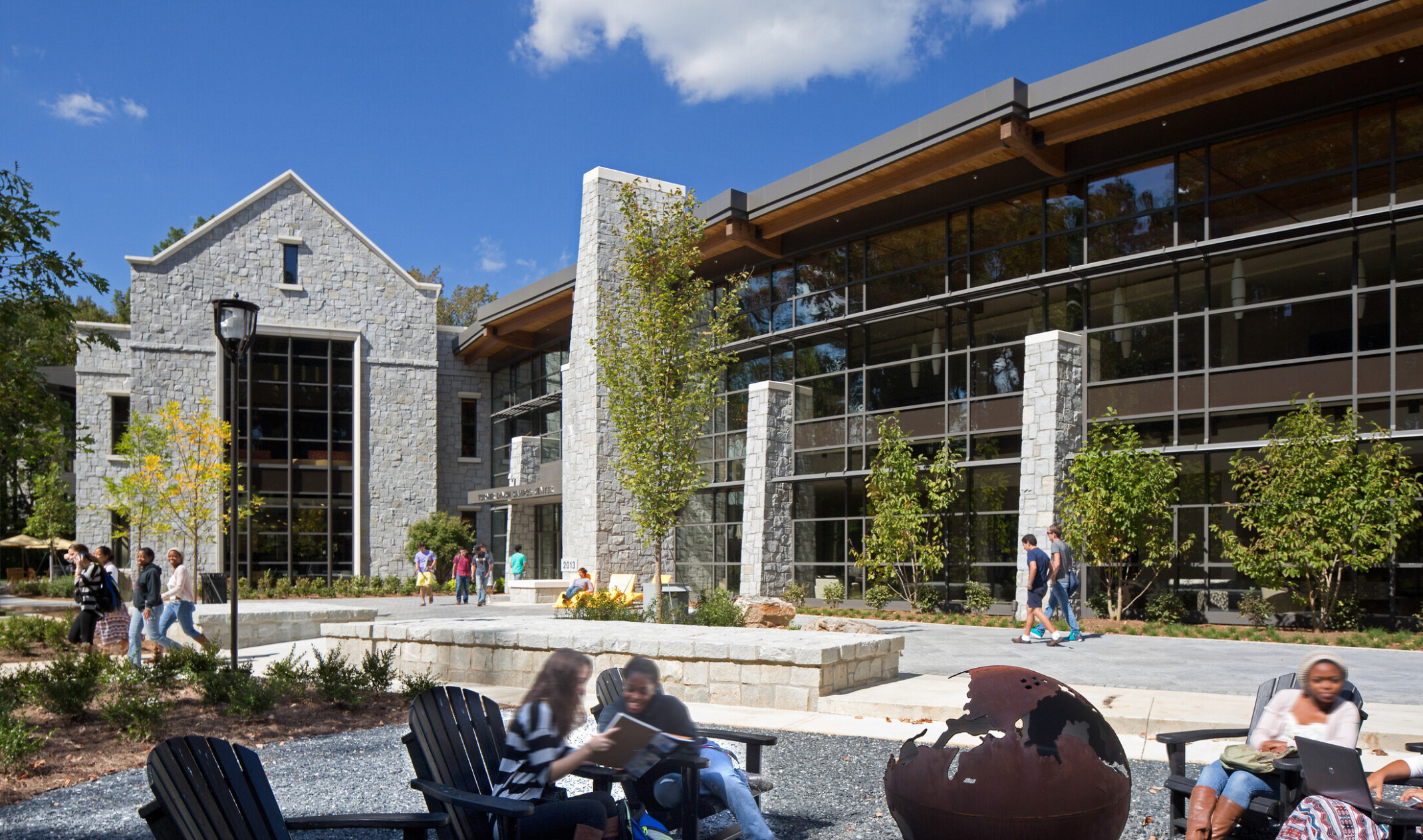 exterior image of a community gathering courtyard; varied seating and greenery surrounded by modern stone and glass building