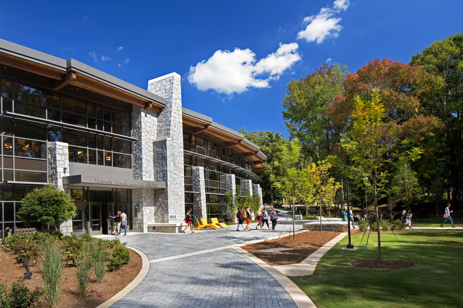 Ogelthorpe Campus Center; curved exposed stone and wood building in front of a gray pathway and green space; students walking in and around building entrance