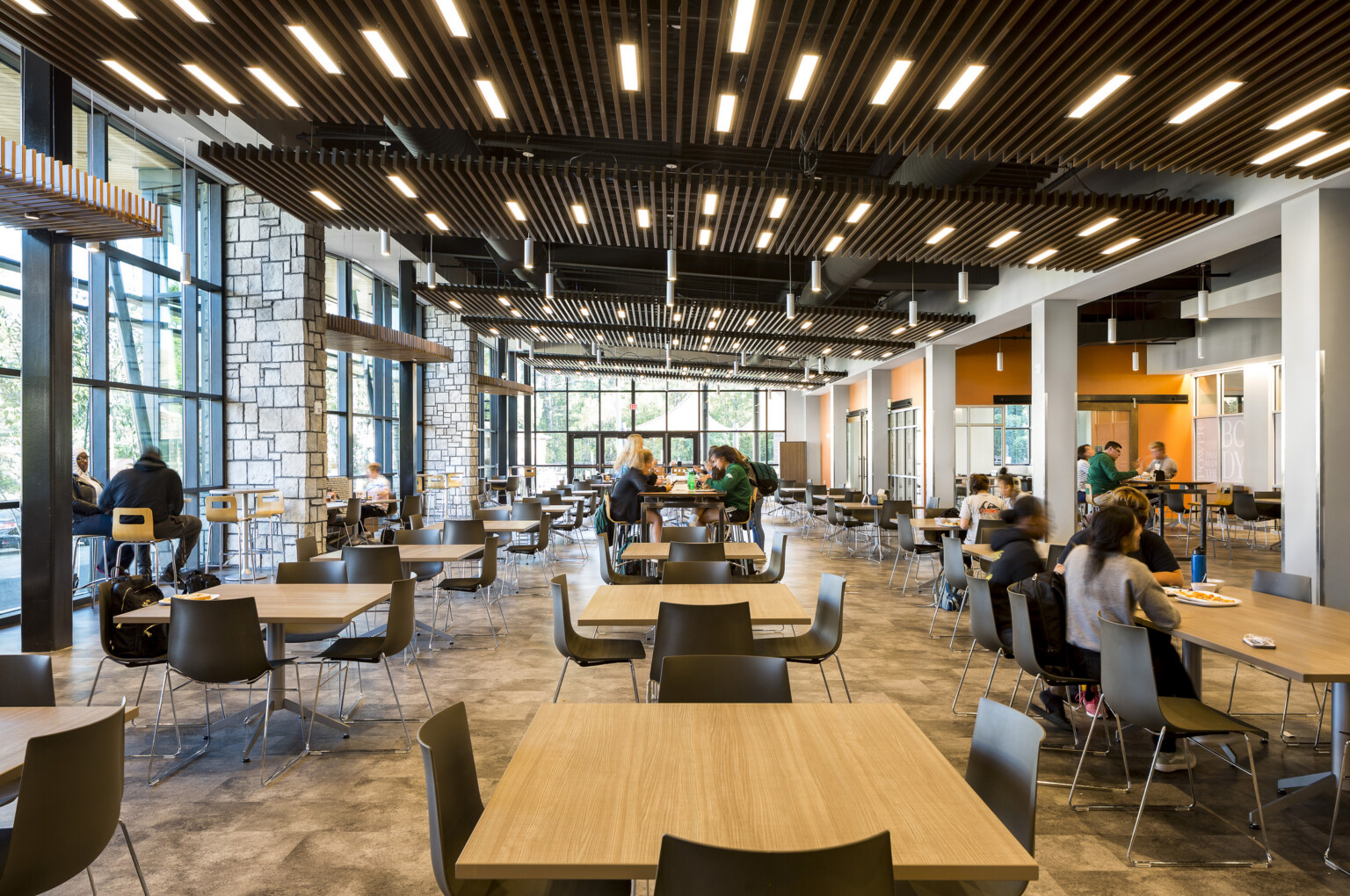 central student hub with tables; students eating and working in the naturally lit space; exposed stone columns and orange accent walls