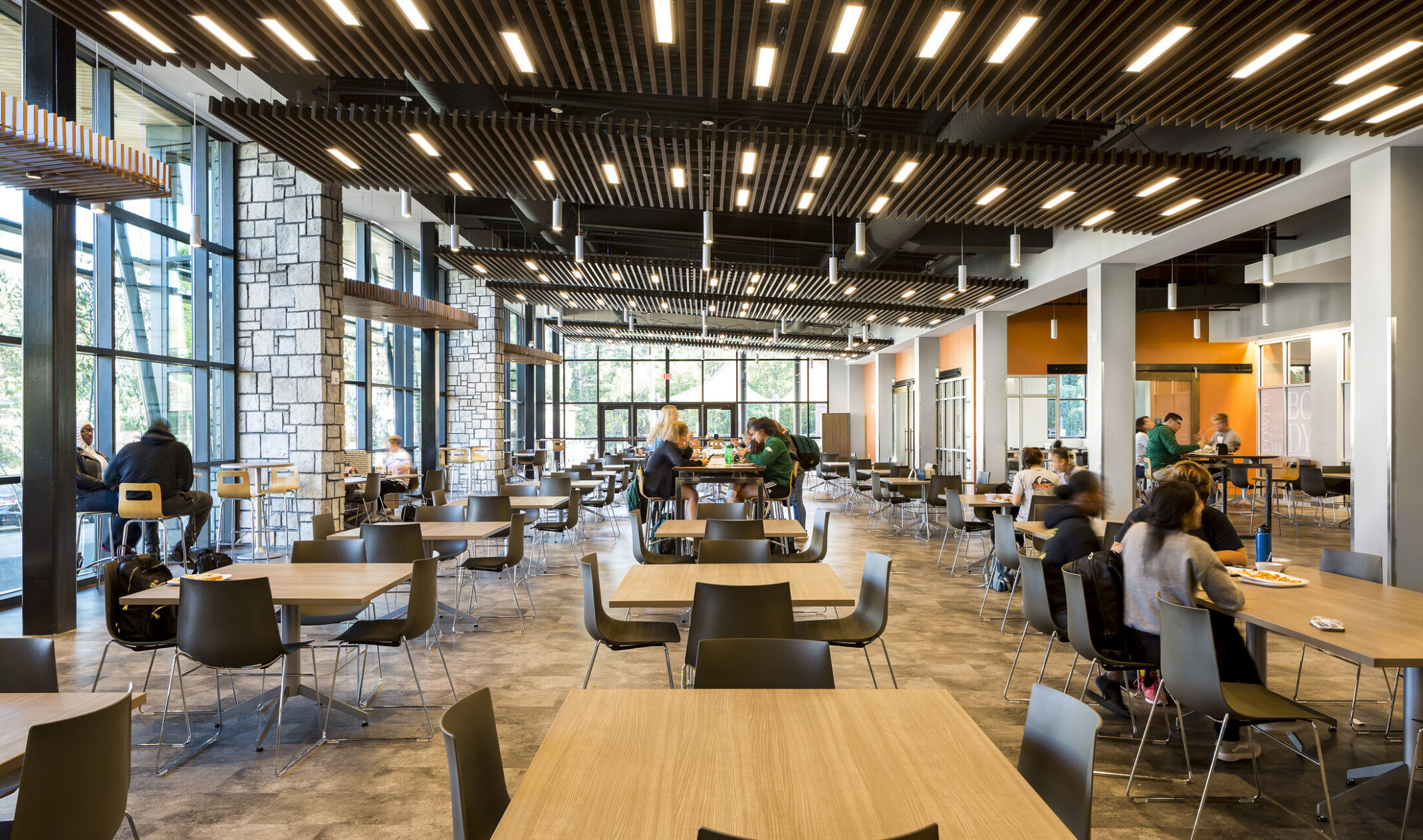 central student hub with tables; students eating and working in the naturally lit space; exposed stone columns and orange accent walls