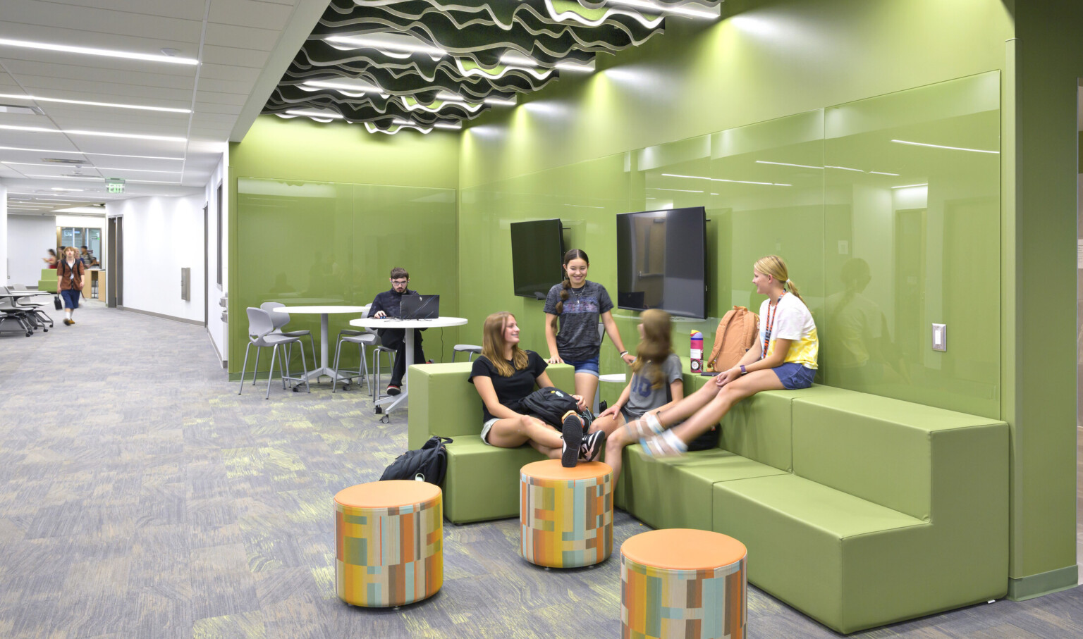 School-aged children sitting in a green collaboration area; bleacher style benches allow for multi-use seating; free standing tables and ottomans for laptops; the green space is lit by an accent ceiling light and embedded in a hallway