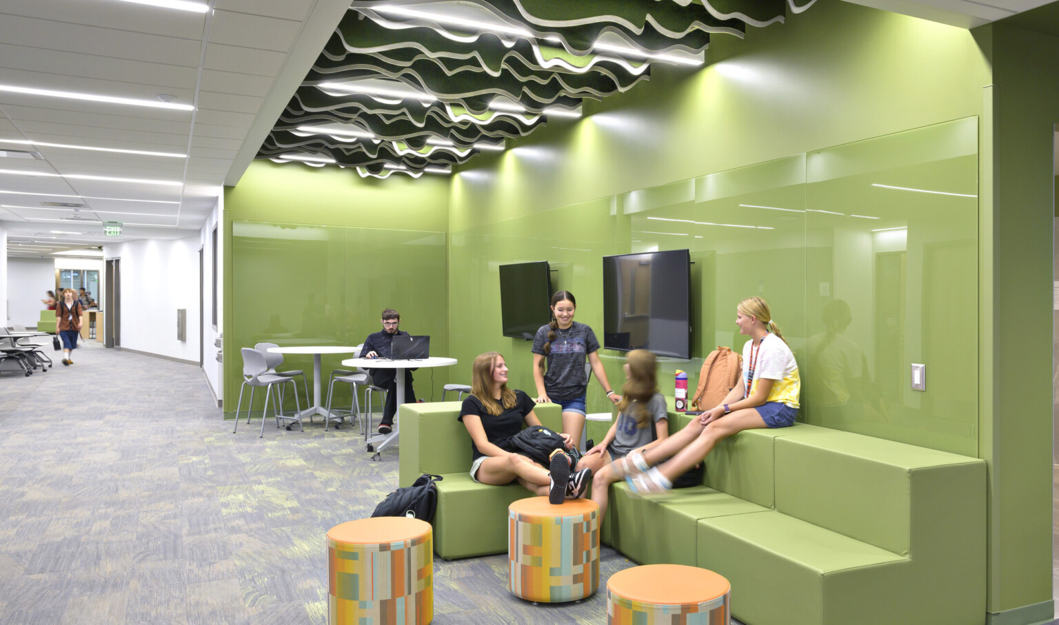 School-aged children sitting in a green collaboration area; bleacher style benches allow for multi-use seating; free standing tables and ottomans for laptops; the green space is lit by an accent ceiling light and embedded in a hallway