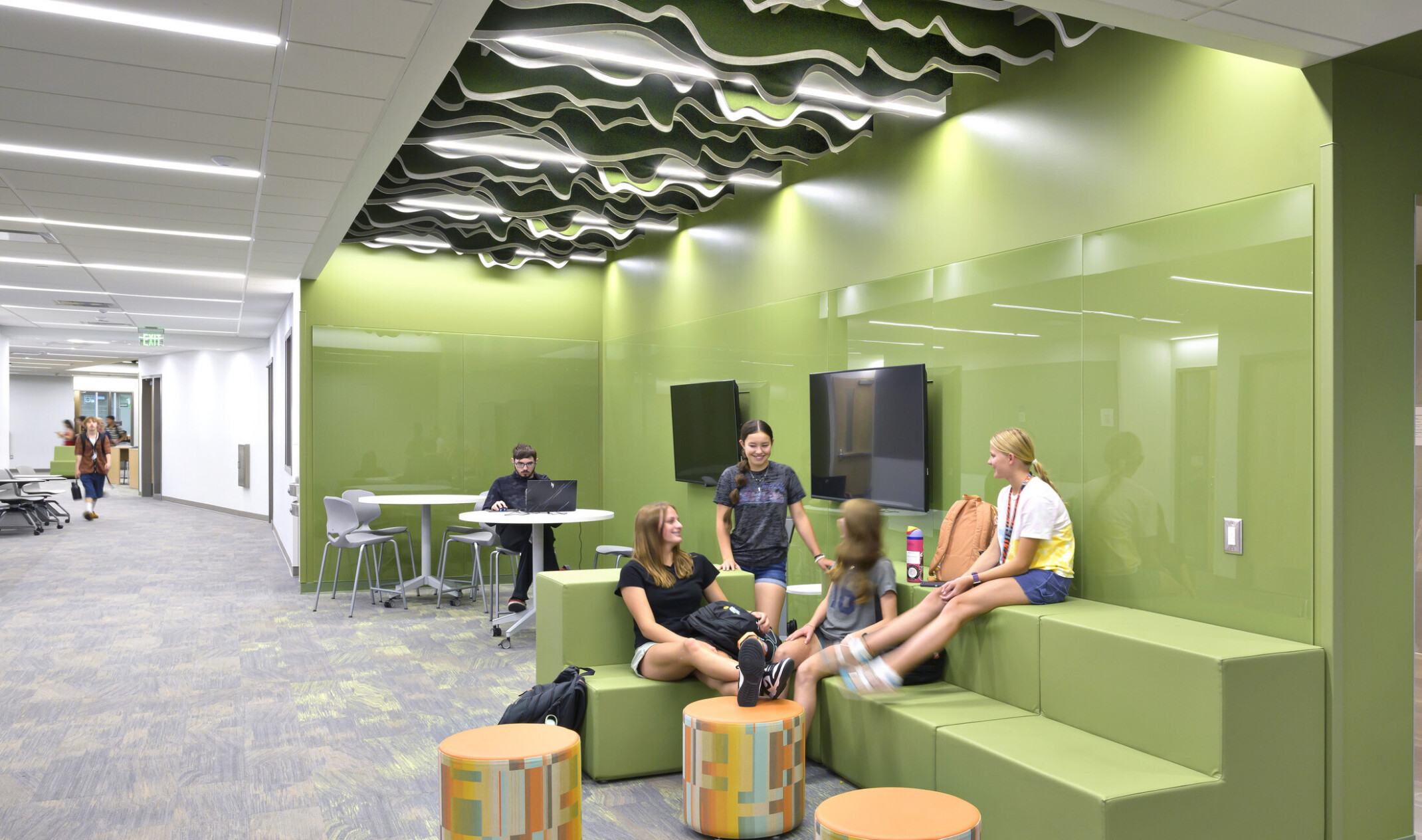 School-aged children sitting in a green collaboration area; bleacher style benches allow for multi-use seating; free standing tables and ottomans for laptops; the green space is lit by an accent ceiling light and embedded in a hallway