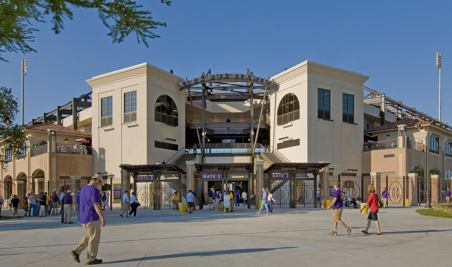 Gated entry labeled Alex Box Stadium Gate 2. Metal fence with baseball design in front of double staircase by white buildings