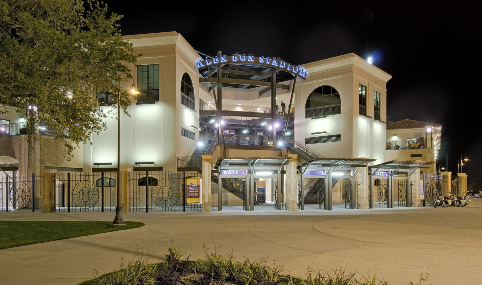 Gate 2 illuminated by fixtures on black exposed beams of stairs, upper walkway, and supports for lit Alex Box Stadium sign